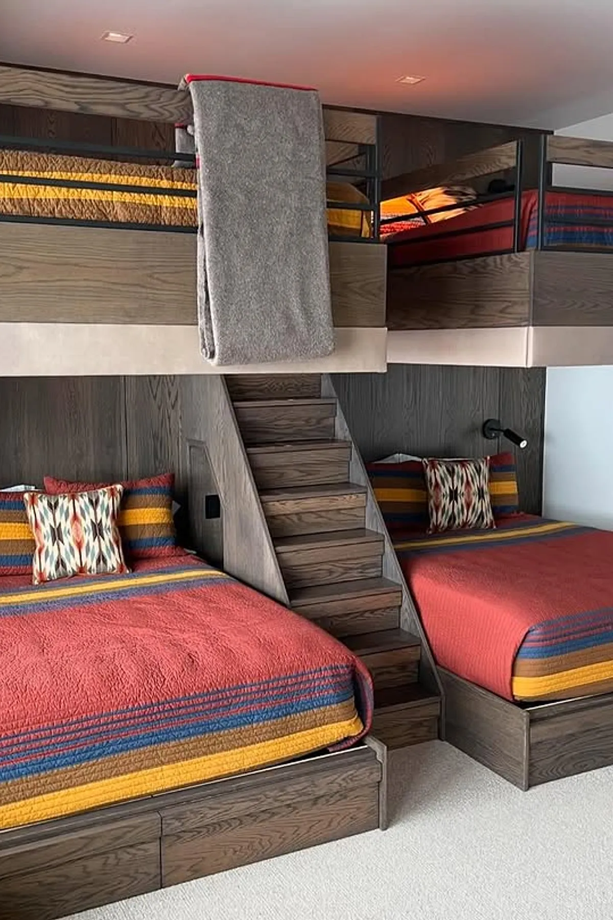 Built-in bunk beds with colorful striped bedding, wooden structure, central staircase, and under-stair storage in a modern rustic room.
