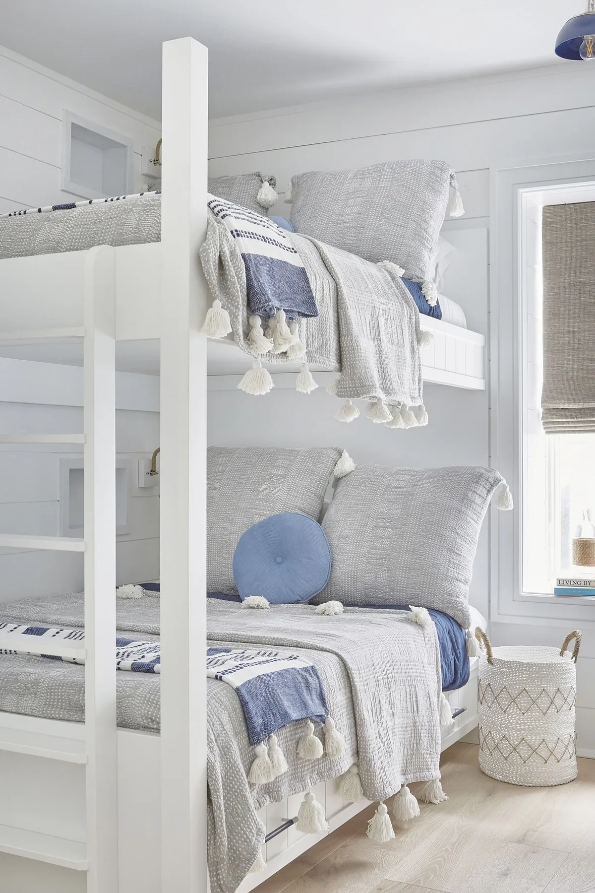 Built-in bunk beds with grey and blue tassel bedding, white frame, and natural light from a window in a nautical-themed room.