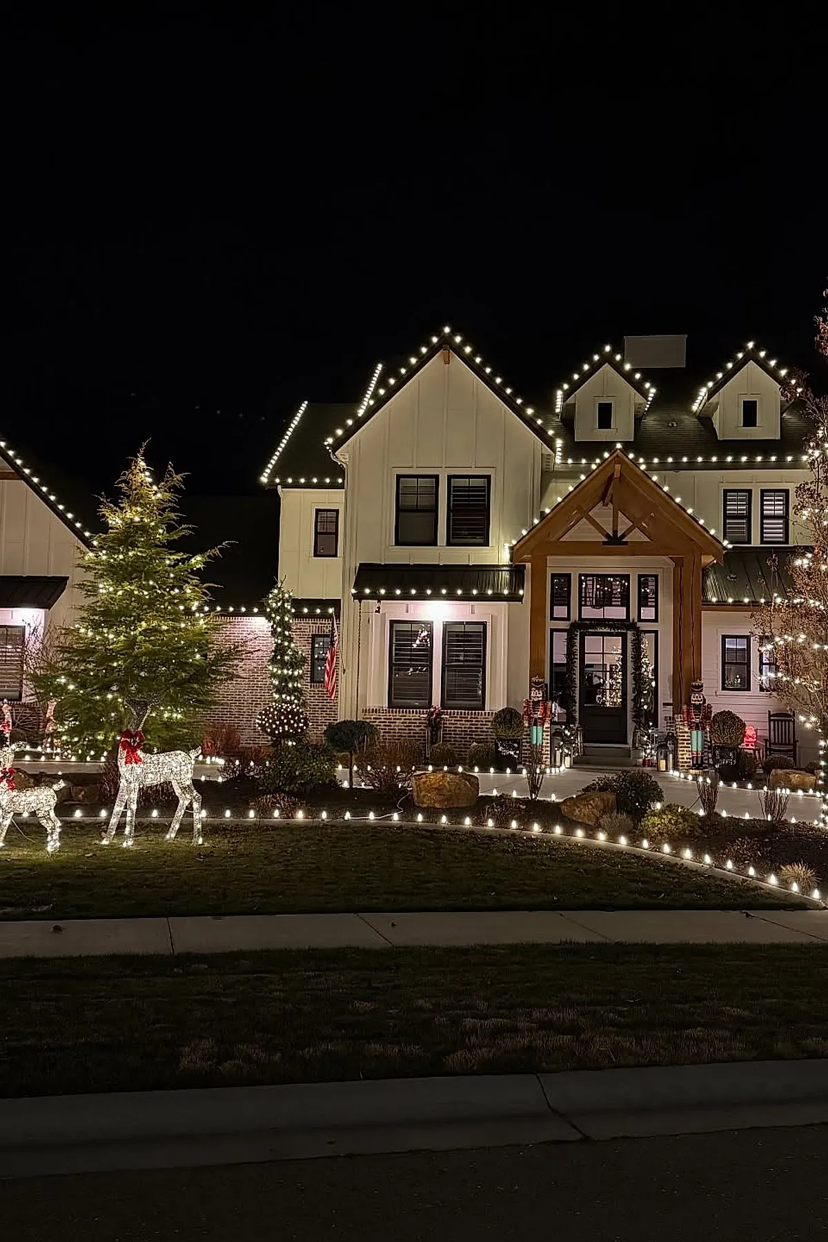White multi-story house with black framed windows decorated with warm white outdoor christmas lights outlining the roof, porch, and landscaping; illuminated reindeer figures with red bows stand on the lawn near a large evergreen tree wrapped in white lights.