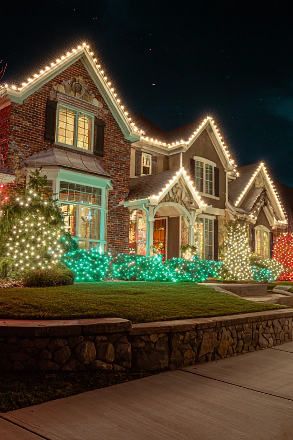 Brick house at night decorated with warm white christmas lights outlining the roof peaks and porch, surrounded by bushes wrapped in multicolored green, red, and white christmas lights creating a vibrant outdoor Christmas light display.