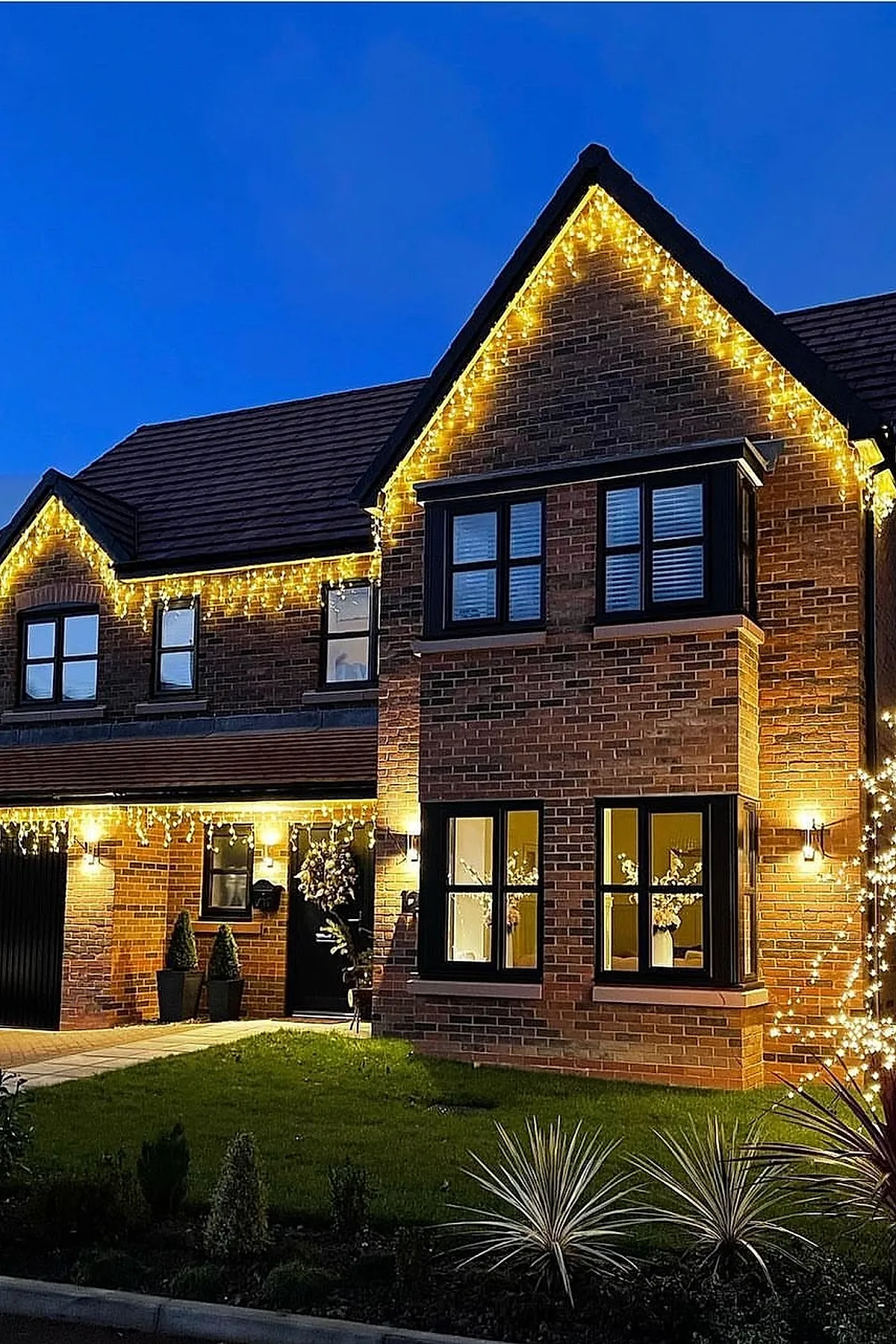 Brick house exterior decorated with warm yellow christmas lights outlining the roof peaks and porch area, illuminating black framed windows and a green lawn under a clear blue evening sky.