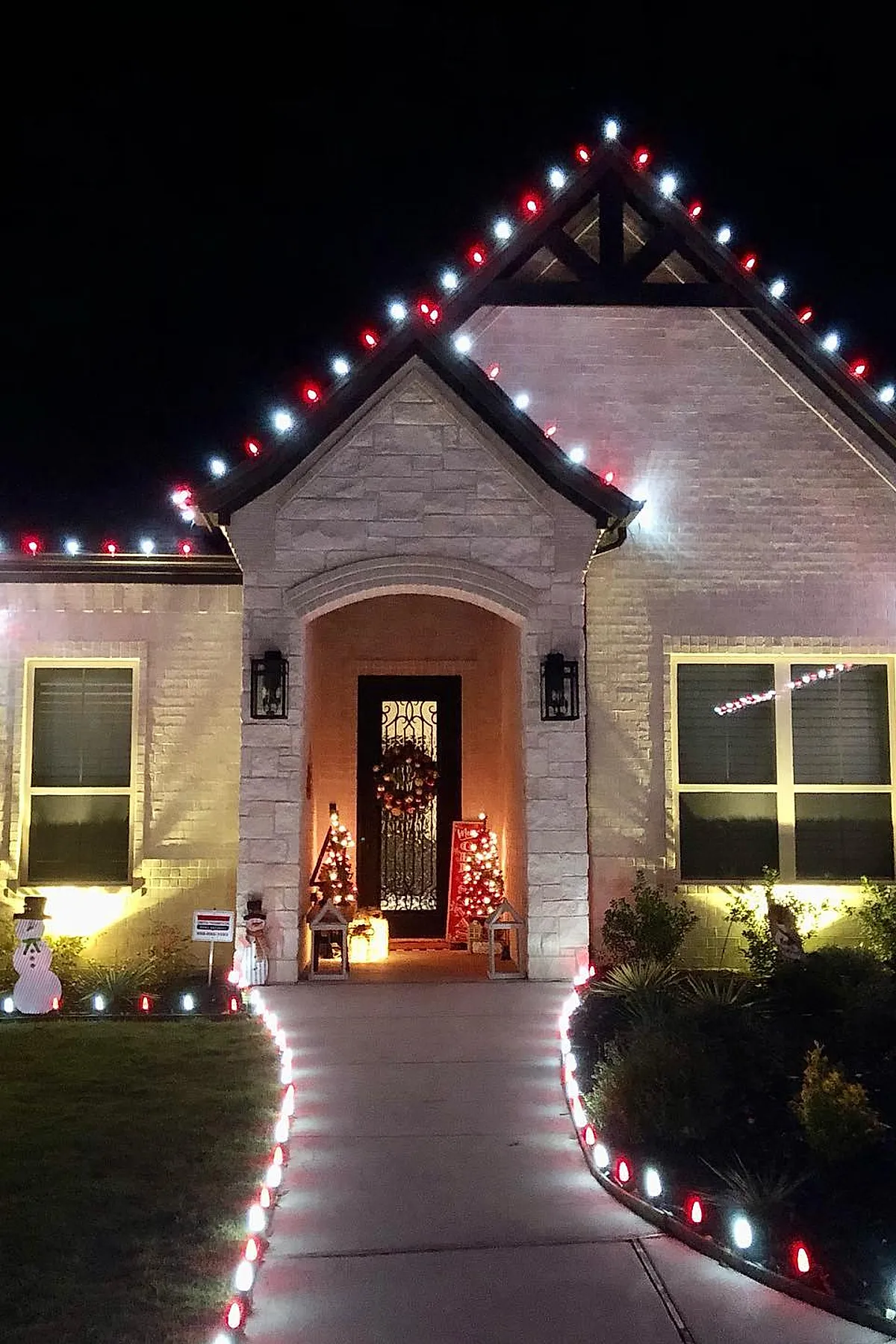 House exterior at night with red and white christmas lights outlining the roofline and lining both sides of the concrete walkway leading to a black front door decorated with a wreath, flanked by illuminated miniature Christmas trees and festive lawn ornaments.
