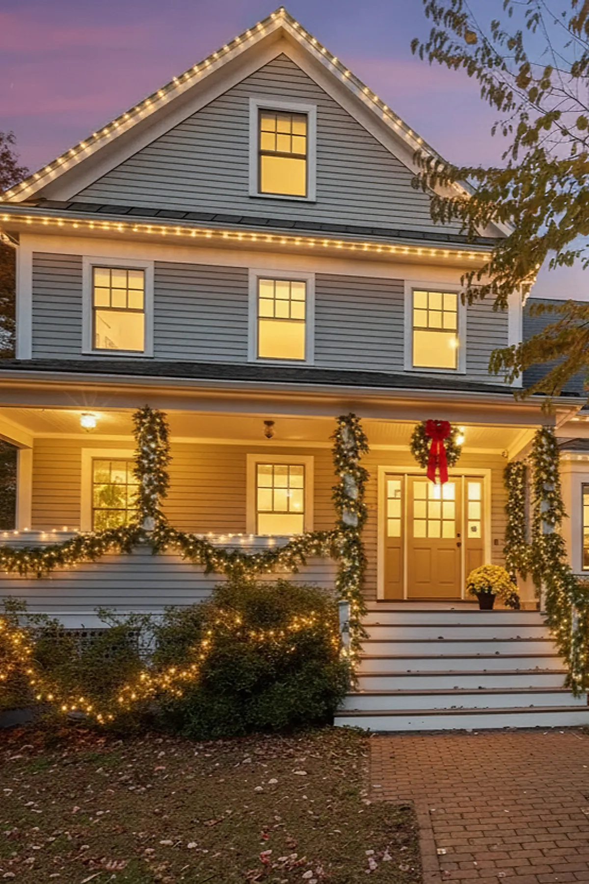 Gray house exterior with warm white christmas lights outlining the roof and porch railings, garlands wrapped around porch columns and railing, a wreath with a large red bow hanging on the front door, all glowing warmly at dusk.