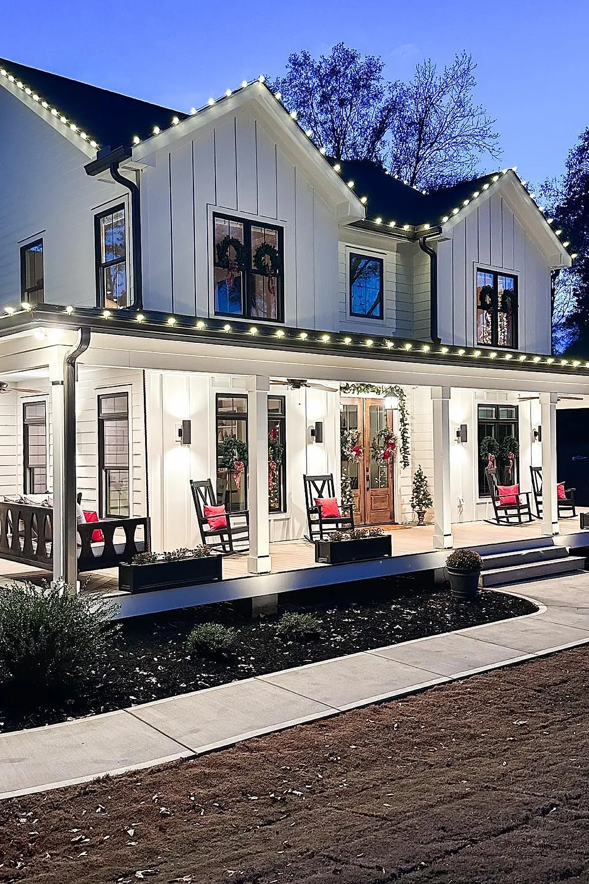 White two-story house with warm white christmas lights outlining the roof and porch, decorated with wreaths on windows and doors, black rocking chairs with red pillows on the porch, and small potted plants.