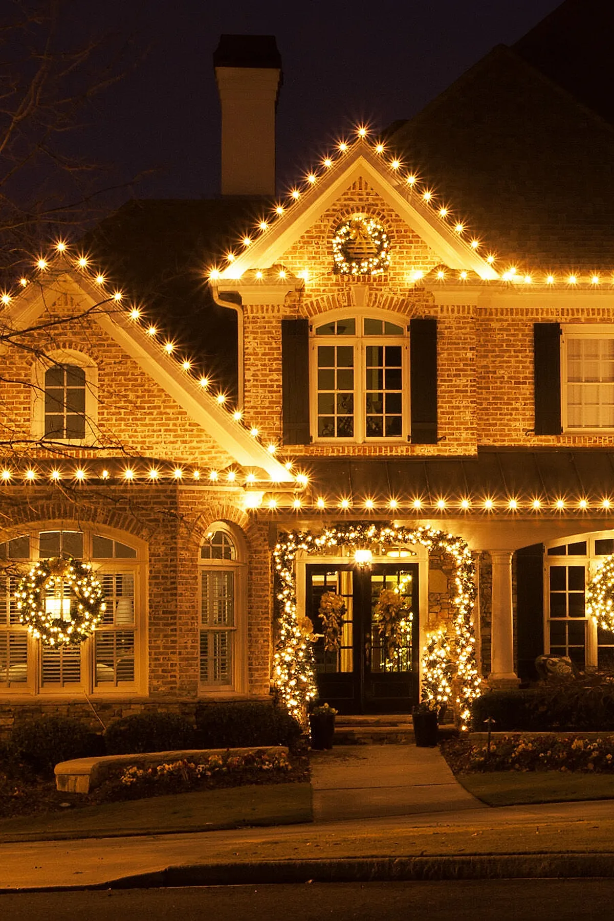 Brick house with warm golden christmas lights outlining roof peaks and porch, illuminated wreaths hanging on windows and above the front door framed by garland wrapped in white lights, set against a dark night sky.