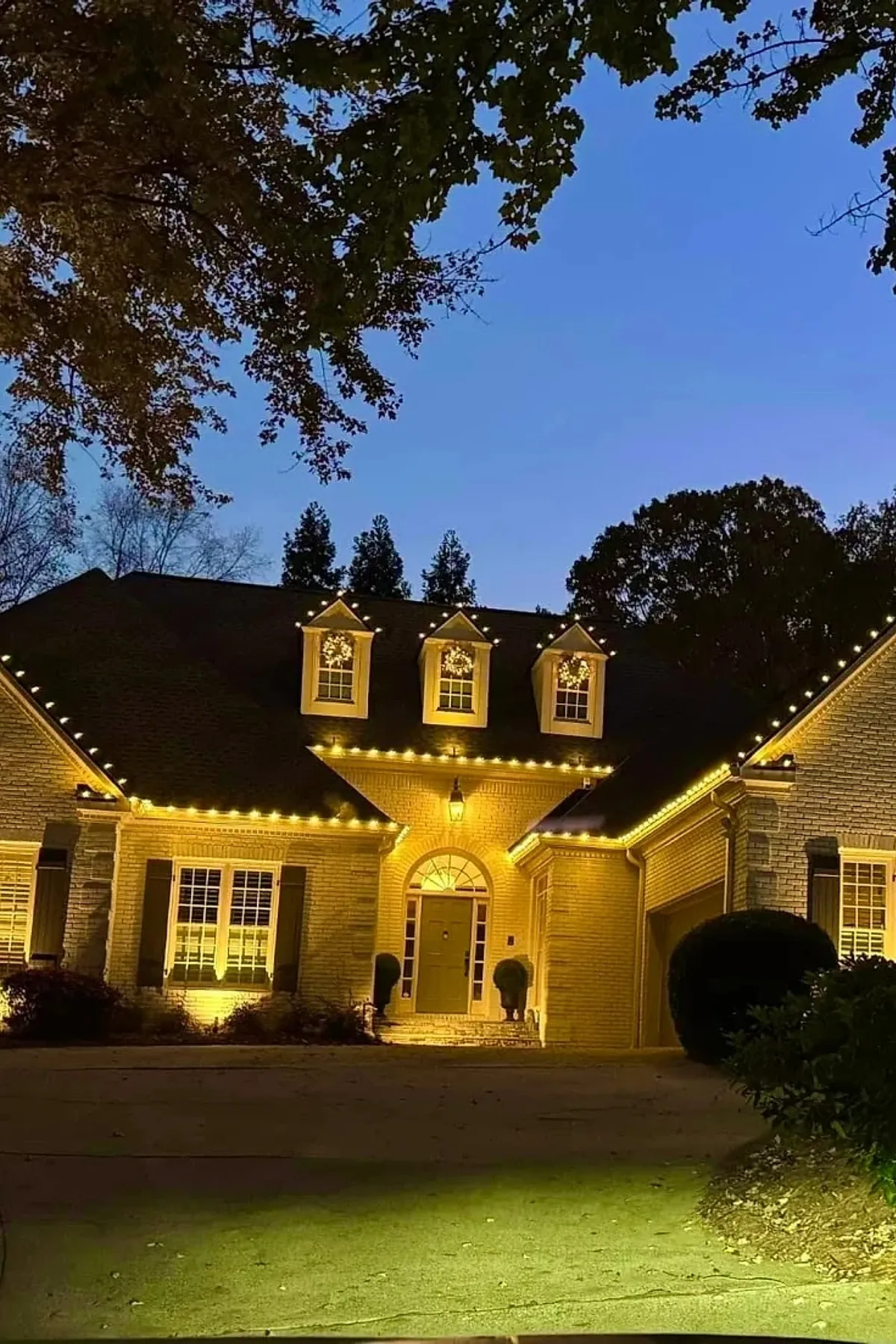 Brick house at dusk with warm yellow christmas lights outlining roof edges and dormer windows, each dormer decorated with illuminated wreaths, creating a warm and inviting outdoor Christmas decoration.
