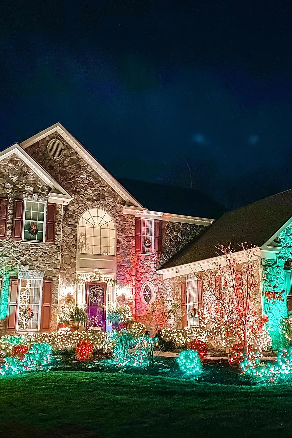 Stone exterior house decorated with bright multicolored outdoor christmas lights in red, green, and white covering bushes and trees around the entrance, with wreaths on windows and a glowing garland framing the front door under a dark night sky.