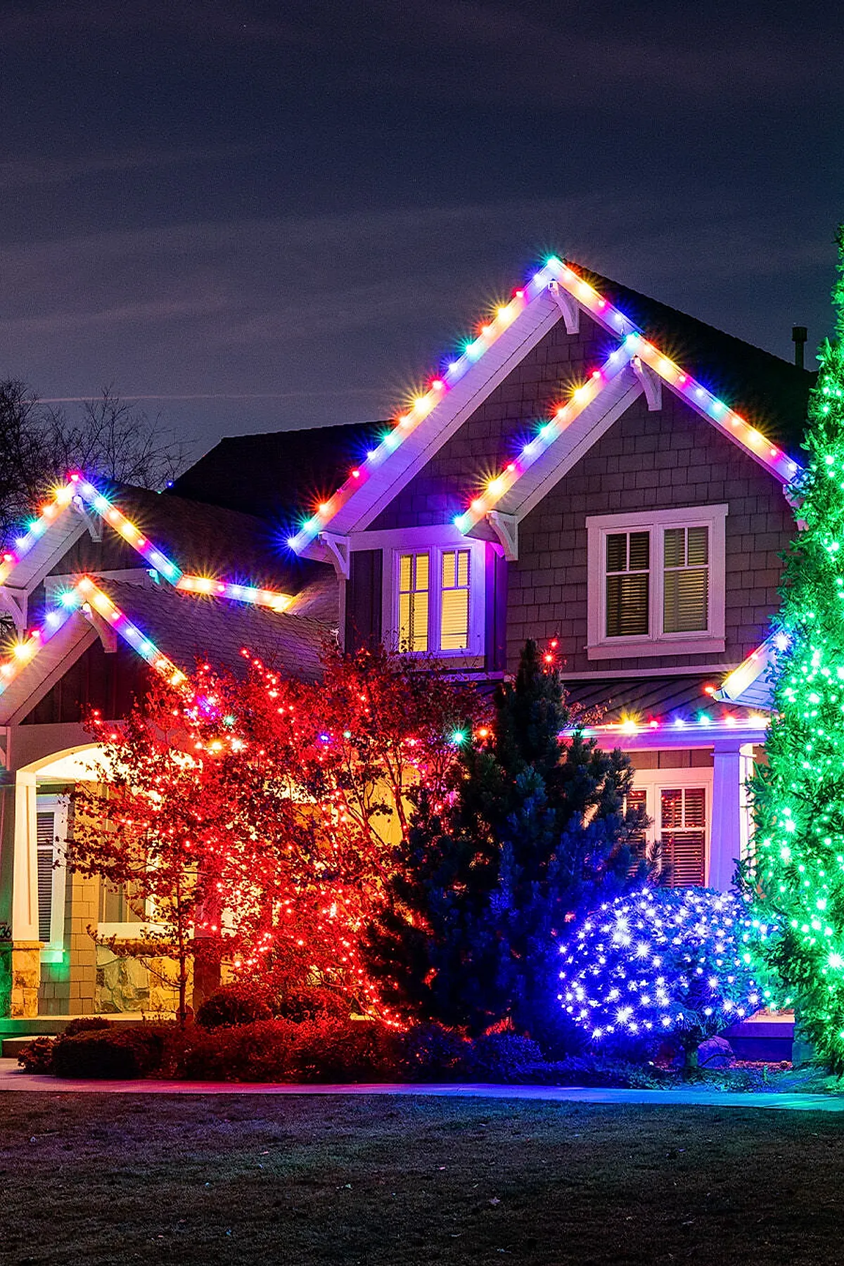 A house decorated with vibrant multicolored christmas lights outlining roof peaks and trim, with bushes and trees wrapped in red, blue, green, and white outdoor christmas lights creating a bright holiday display at night.
