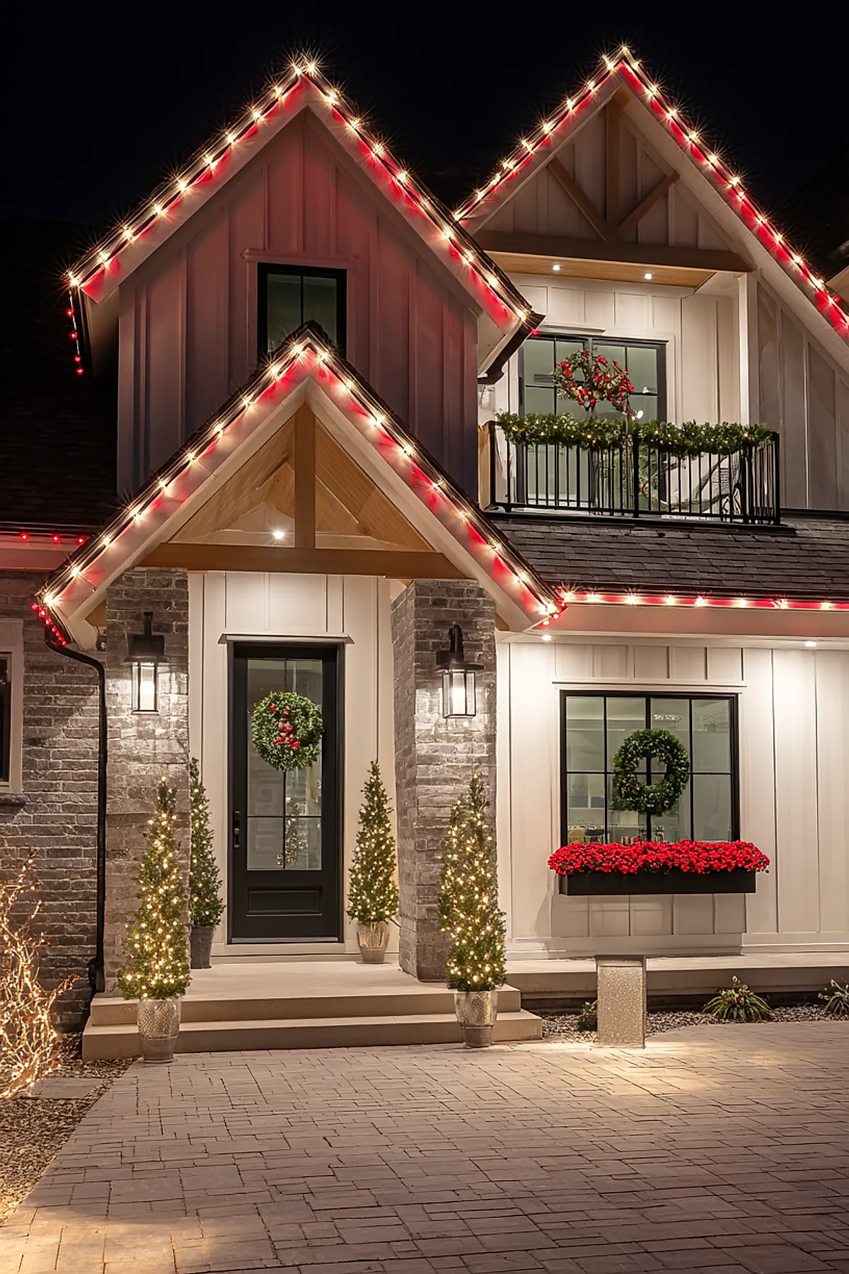 Modern house exterior at night decorated with warm white and red christmas lights outlining roof peaks and porch, featuring a black front door adorned with a green wreath with red berries, flanked by small lit Christmas trees in pots, a balcony decorated with green garland and a wreath, and a flower box filled with red flowers beneath a window.
