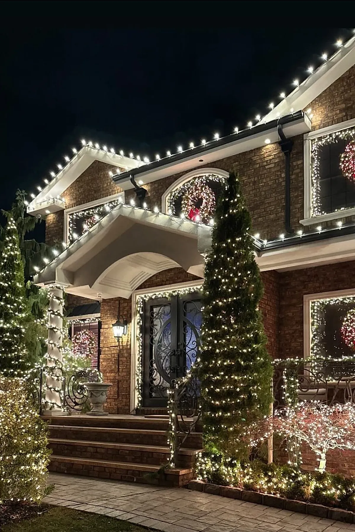 Brick house exterior decorated with warm white outdoor christmas lights outlining roof edges, windows, porch columns, railings, and bushes; wreaths with red bows are illuminated in windows under a dark night sky.