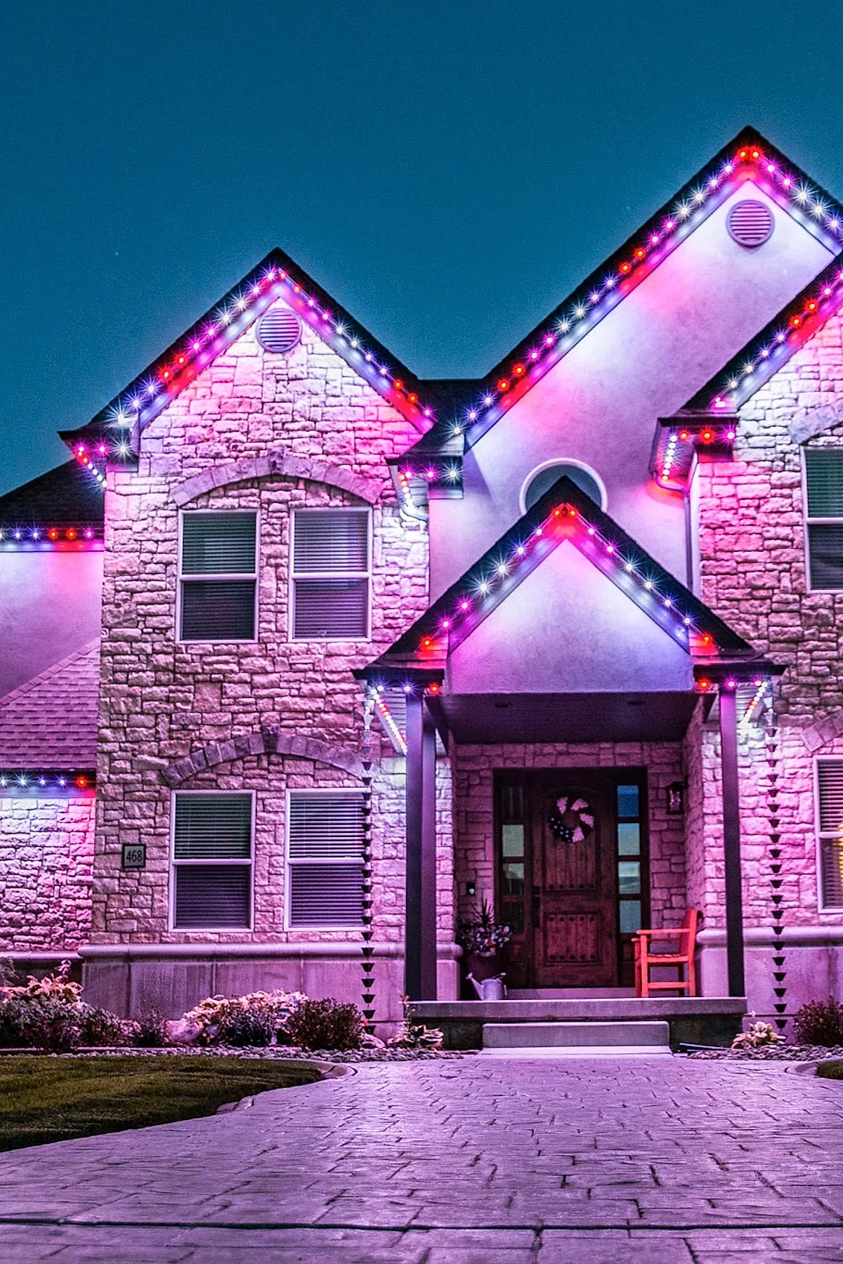 Stone and stucco house exterior decorated with bright pink, purple, red, and white outdoor christmas lights outlining roof peaks and porch at night, casting a colorful glow on the facade with a wooden front door adorned with a wreath.