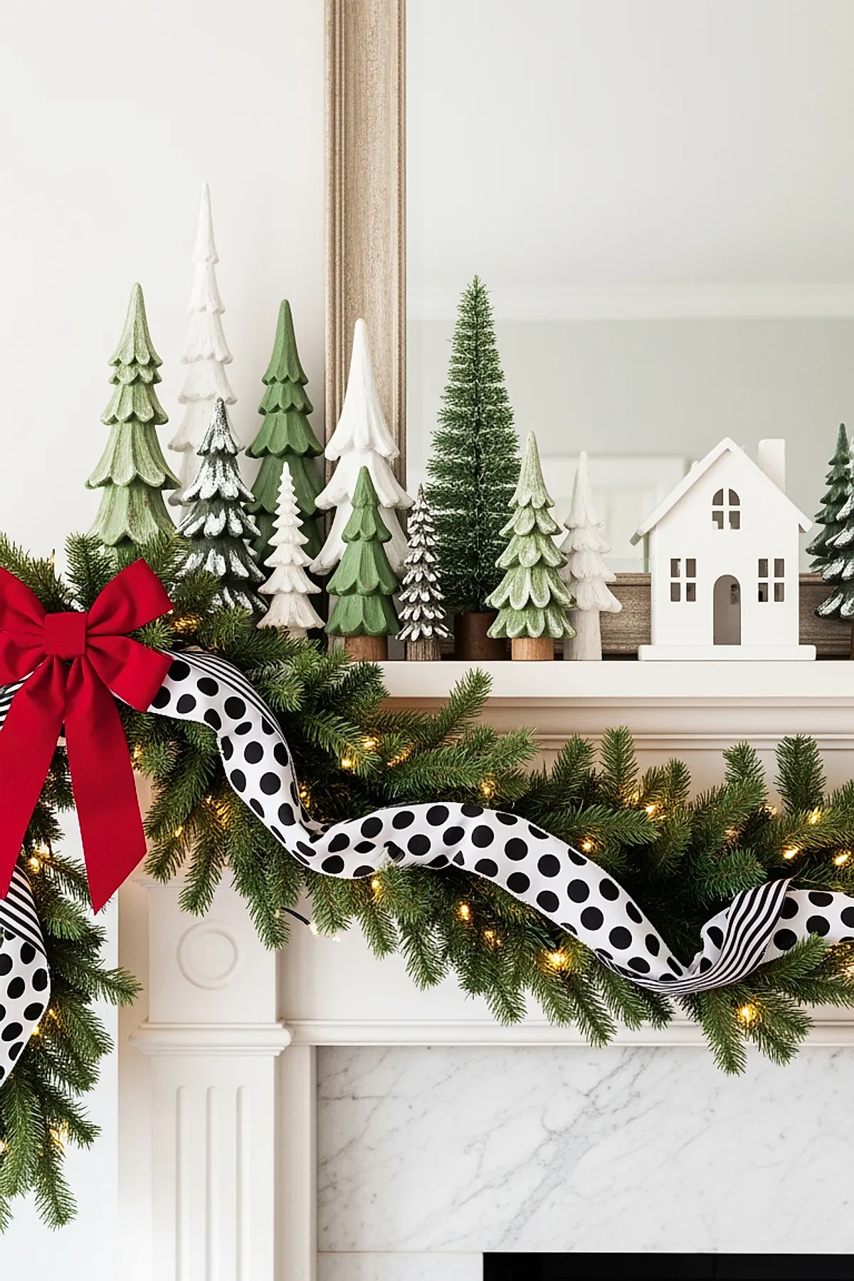 white fireplace mantle decorated with lush green garland wrapped in a black and white polka dot ribbon, accented by a large red bow, topped with various miniature green and white Christmas trees made of wood and bottle brush, alongside a small white decorative house, all set against a large framed mirror
