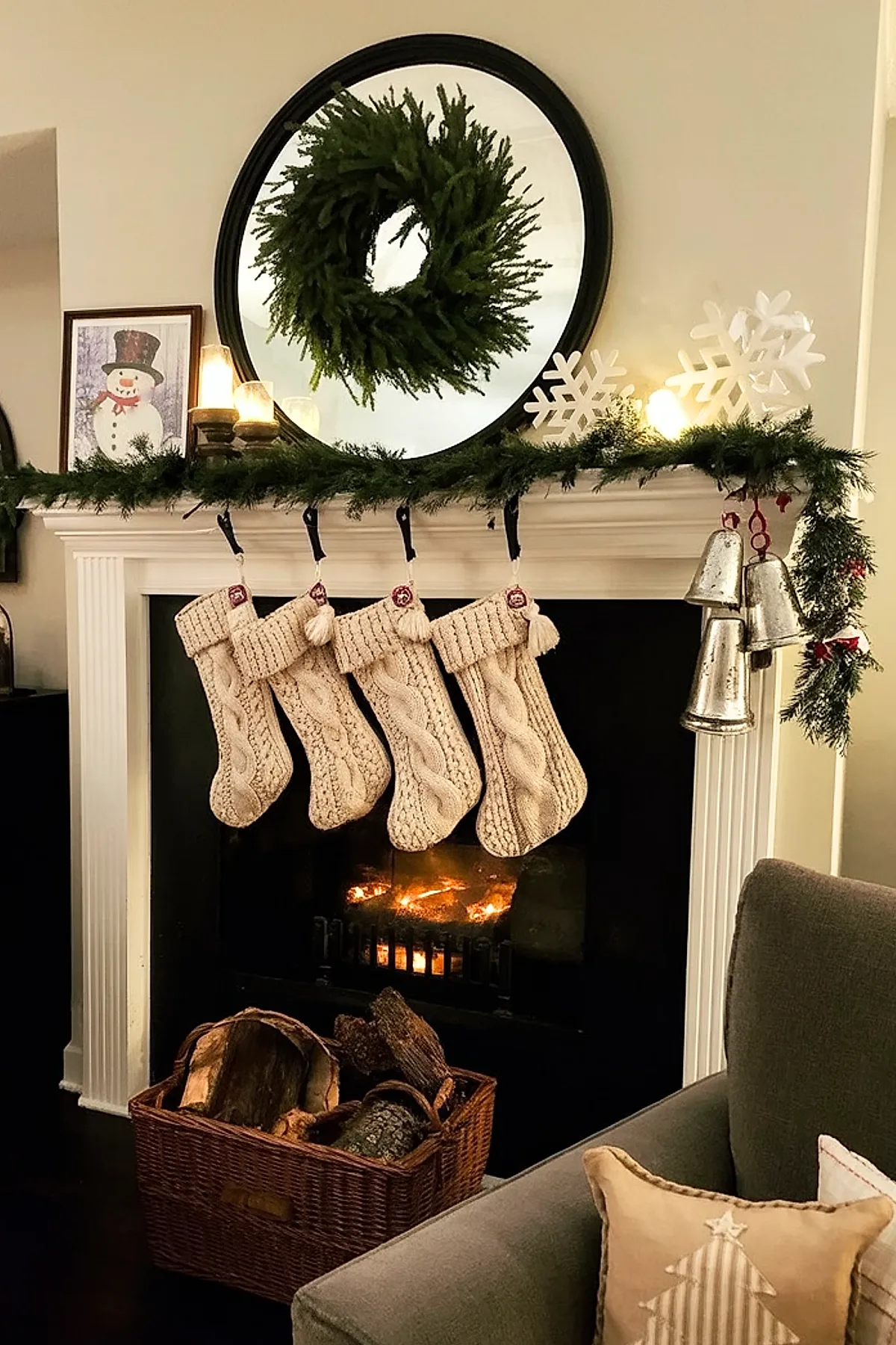 white fireplace mantel decorated with five cream-colored knitted Christmas stockings hanging from black hooks, a lush green wreath reflected in a round black-framed mirror above, greenery garland draped along the top with silver bells hanging on the right side, lit candles and white snowflake decorations on the mantel