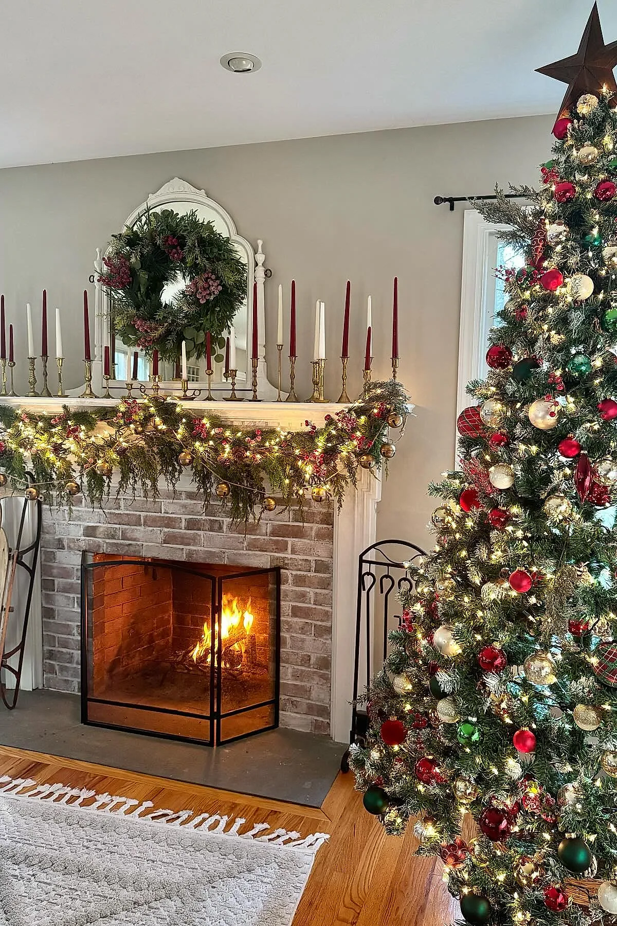 christmas fireplace mantel decorated with a lush green wreath hanging on an ornate white mirror above, red and white taper candles in gold candlesticks arranged symmetrically, greenery garland with string lights and gold ornaments draped across a white wooden mantel, burning fire inside brick fireplace next to a decorated christmas tree with red, gold, and green ornaments topped with a brown star