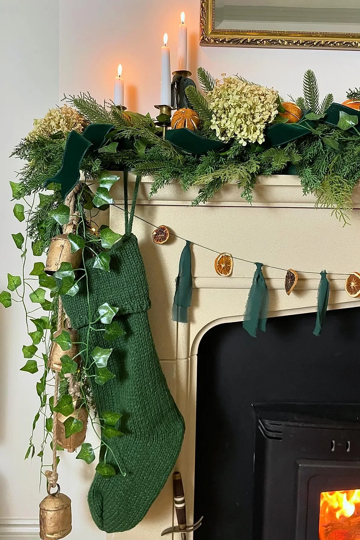 christmas mantle decorated with green knit stocking, lush green garland accented with dried orange slices and light yellow hydrangeas, brass candle holders with white candles, hanging large brass bells, under a gold-framed mirror above a cream fireplace