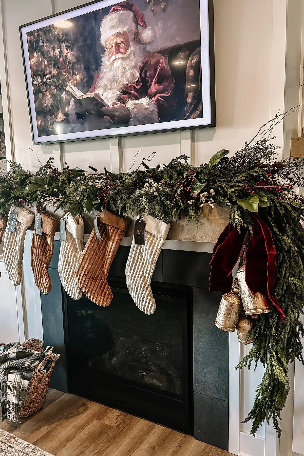 christmas mantle decorated with neutral-toned velvet stockings hanging from a wooden mantel draped in lush green garland accented with white and red berries, gold bells tied with dark red velvet ribbon, under a large framed Santa Claus painting above black fireplace