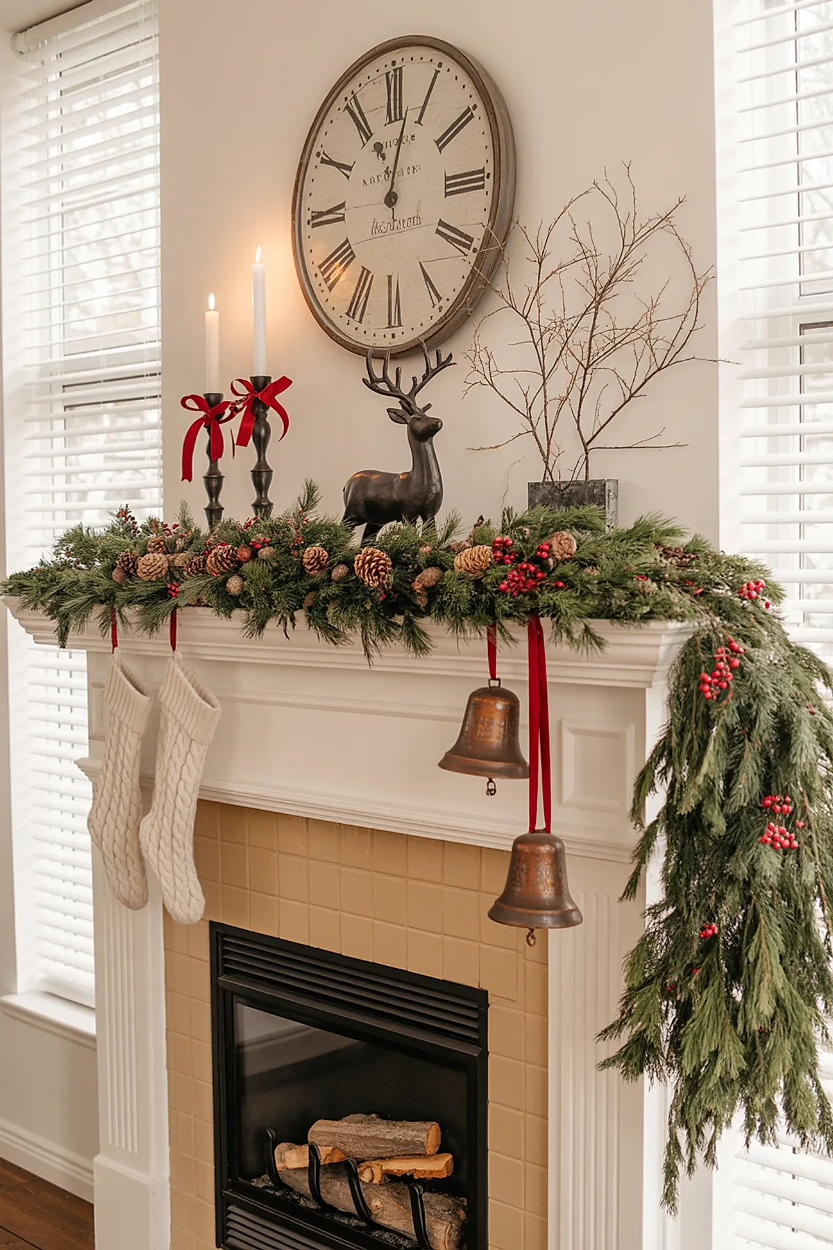 white fireplace mantel decorated with lush green garland featuring pinecones and red berries, two cream knit stockings hanging, two large rusty metal bells tied with red ribbons hanging from the mantel, bronze deer figurine, black candle holders with white taper candles adorned with red ribbon bows, beige tiled fireplace surround