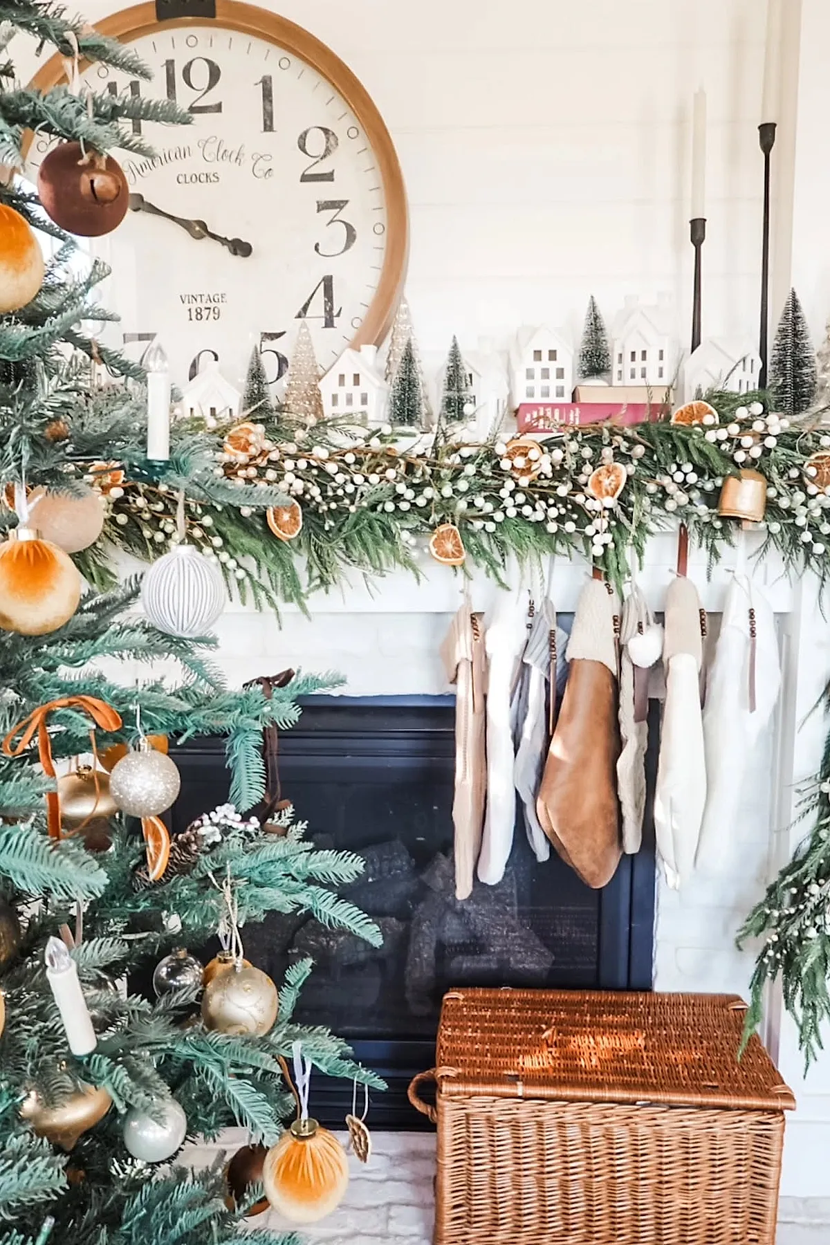 christmas mantle decorated with lush green garland accented with white berries and dried orange slices, hanging neutral-toned stockings, miniature white village houses and small bottle brush trees on top, large vintage-style clock behind, next to a decorated christmas tree with gold and cream ornaments