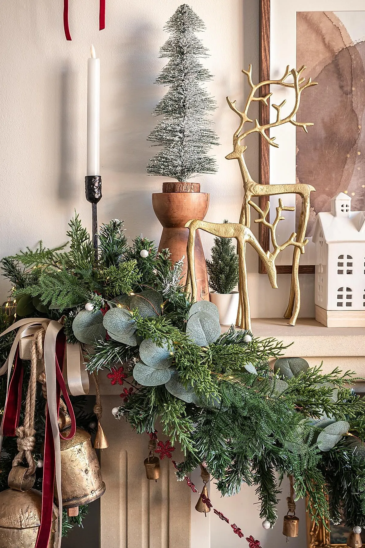 christmas mantle decorated with lush green garland featuring eucalyptus and pine branches, gold metal reindeer figurines in two sizes, small frosted bottle brush tree on a wooden pedestal, white taper candle in black holder, and a white ceramic house against a beige wall.