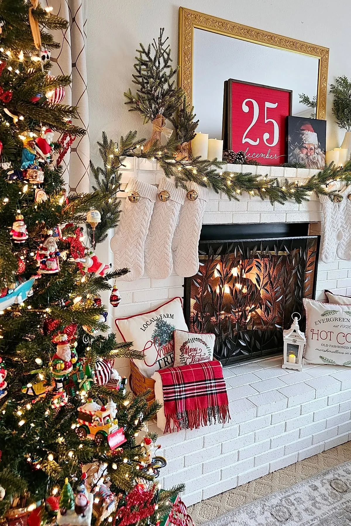 white brick fireplace with white knit stockings hanging from a greenery garland decorated with string lights and jingle bells, red plaid blanket and festive pillows arranged on hearth next to lit fire and Christmas tree with colorful ornaments
