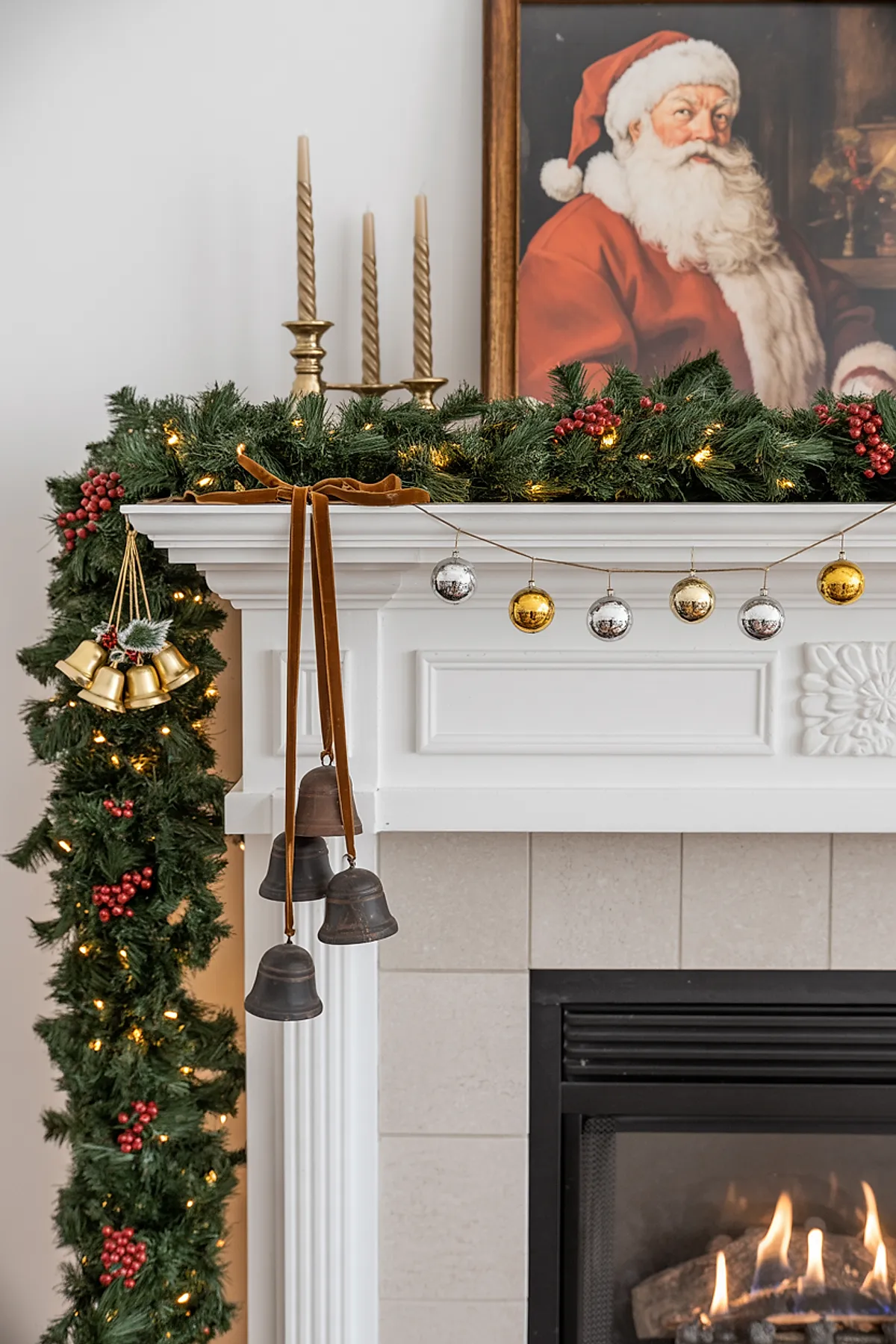 white fireplace mantle decorated with lush green garland featuring red berries and warm string lights, hanging gold and dark metal bells tied with brown velvet ribbons on the left side, gold candlesticks with twisted beige candles on top, a framed vintage Santa portrait above