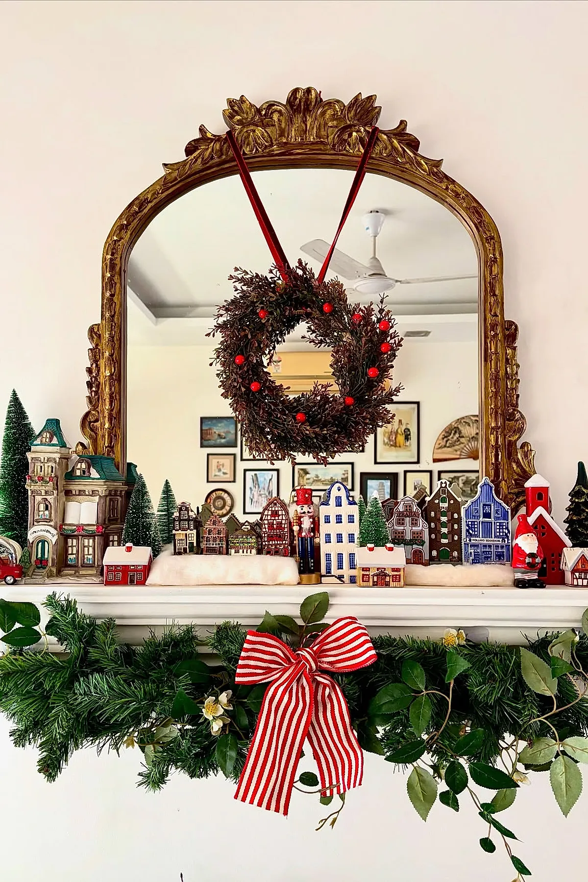 ornate gold-framed mirror above white fireplace mantel decorated with miniature ceramic Christmas village houses, small green pine trees, a nutcracker figure, and a wreath hanging on the mirror with red berries and velvet ribbons; lush green garland with leafy branches and a red and white striped bow drapes across the front of the mantel