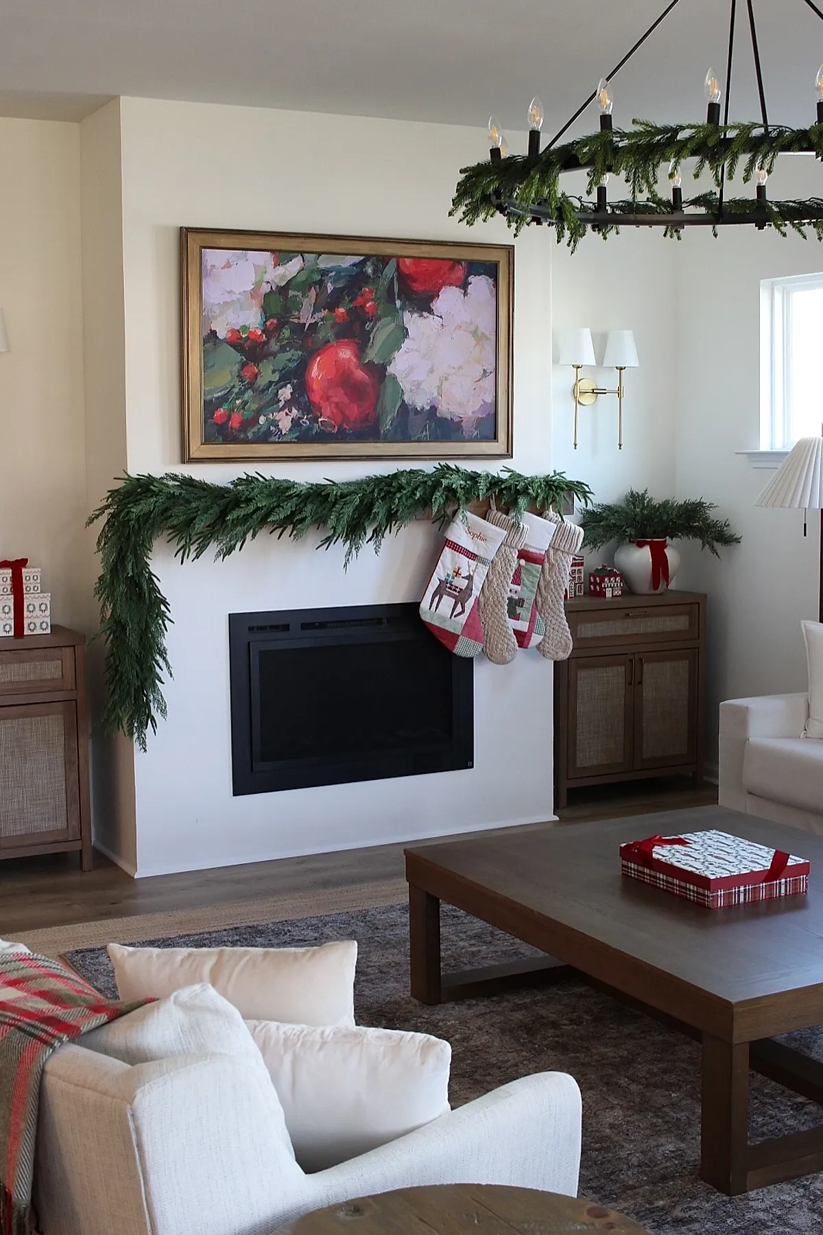 modern white fireplace mantel decorated with a simple lush green garland and three Christmas stockings hanging, beige walls with gold framed floral painting above, wooden sideboards on either side with wrapped gifts and greenery in white vase