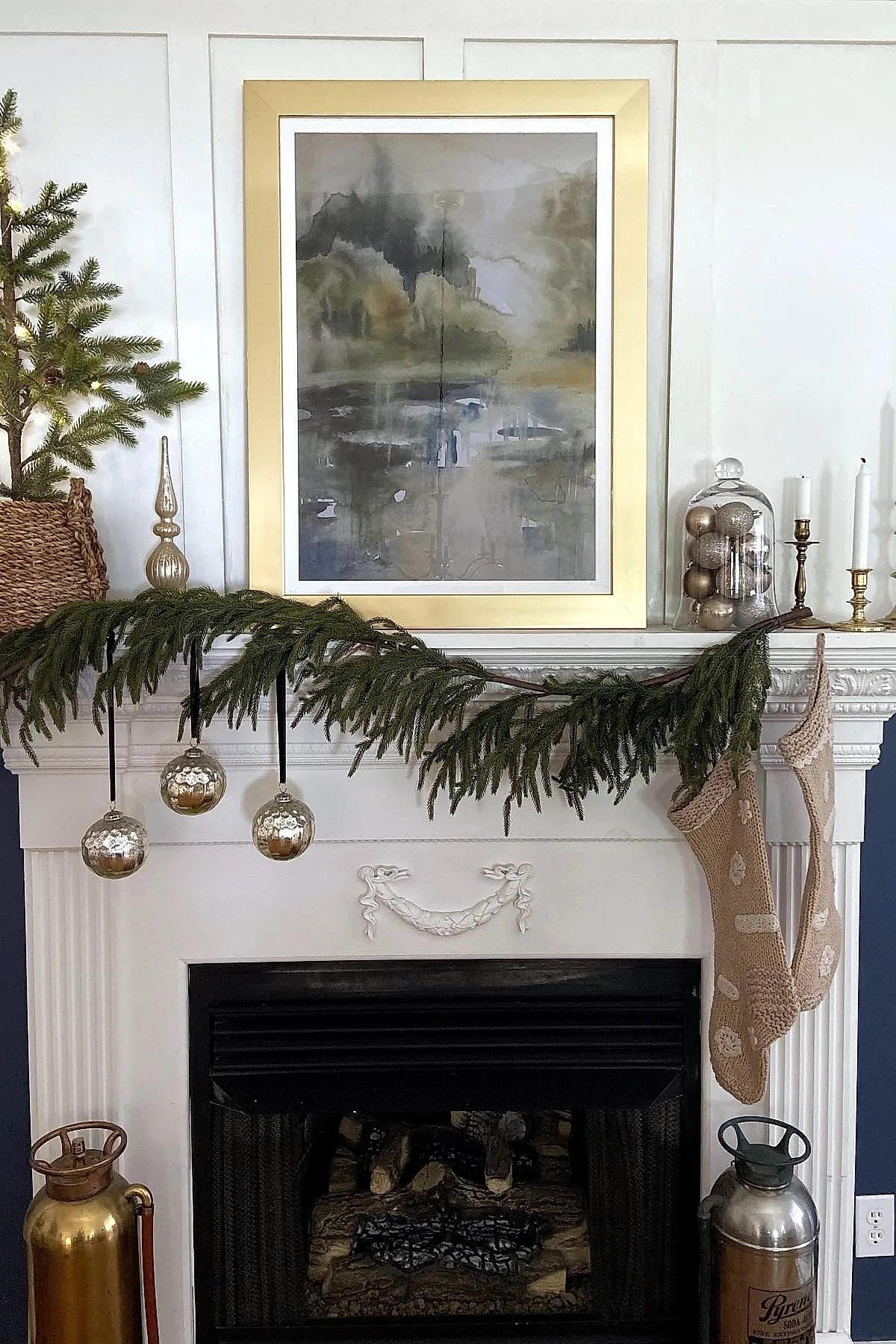 white fireplace mantel decorated with a simple evergreen garland draped across it, three metallic silver Christmas ornaments hanging from black ribbons, two beige knit stockings with white patterns hanging on the right side, brass candlesticks and glass cloche filled with silver ornaments on top, set against white paneled walls and a large abstract painting in gold frame above
