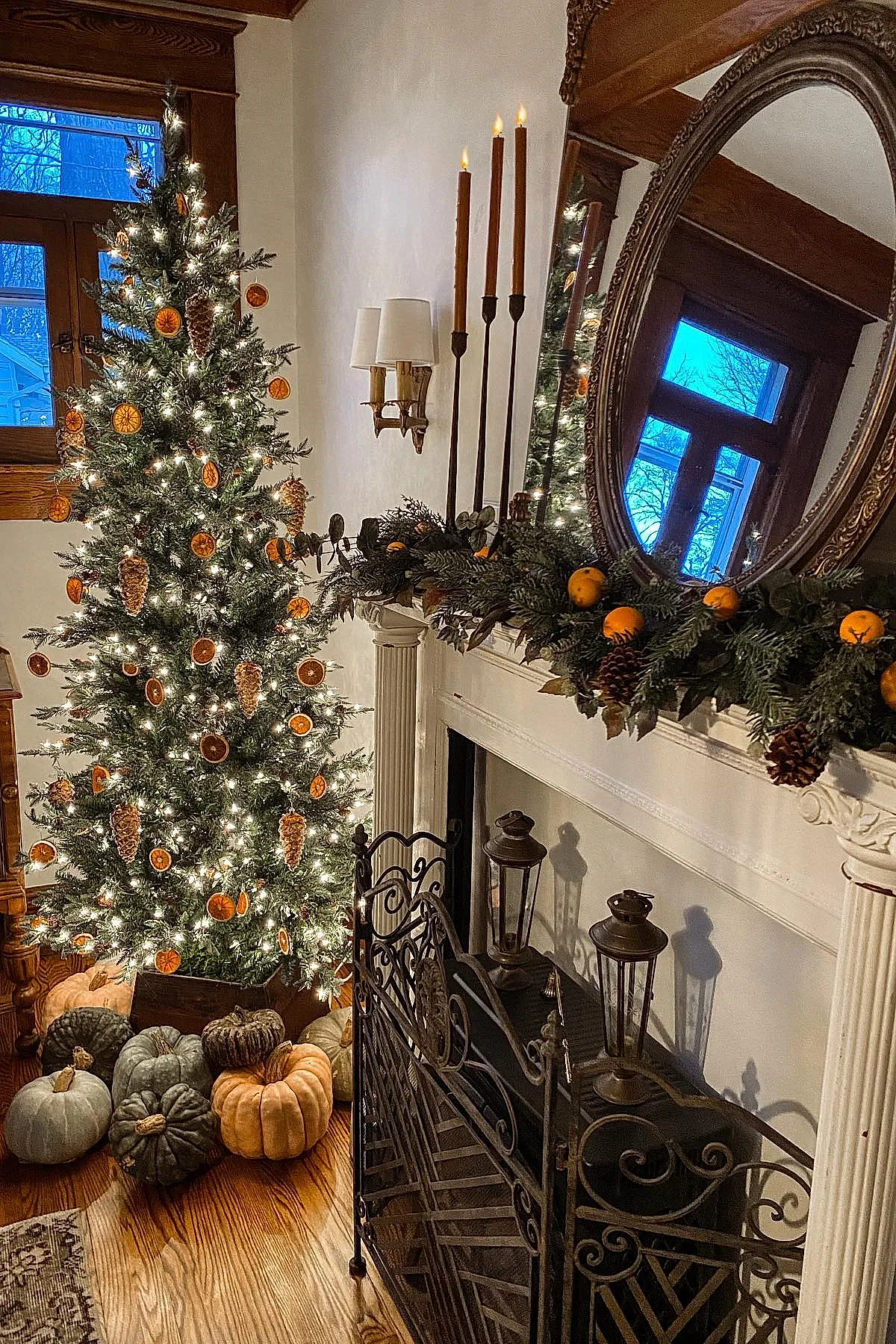 christmas mantle decorated with greenery garland featuring pinecones and dried orange slices, tall brown taper candles in black holders, an ornate round mirror above white fireplace, with a lit Christmas tree adorned with similar dried orange ornaments beside pumpkins on wooden floor