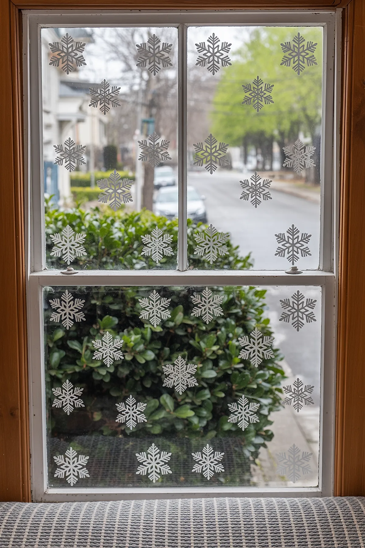 Christmas window art featuring evenly spaced white snowflake cling decals across four glass panes framed by wooden trim, with greenery and a street view outside for an easy DIY winter window display.