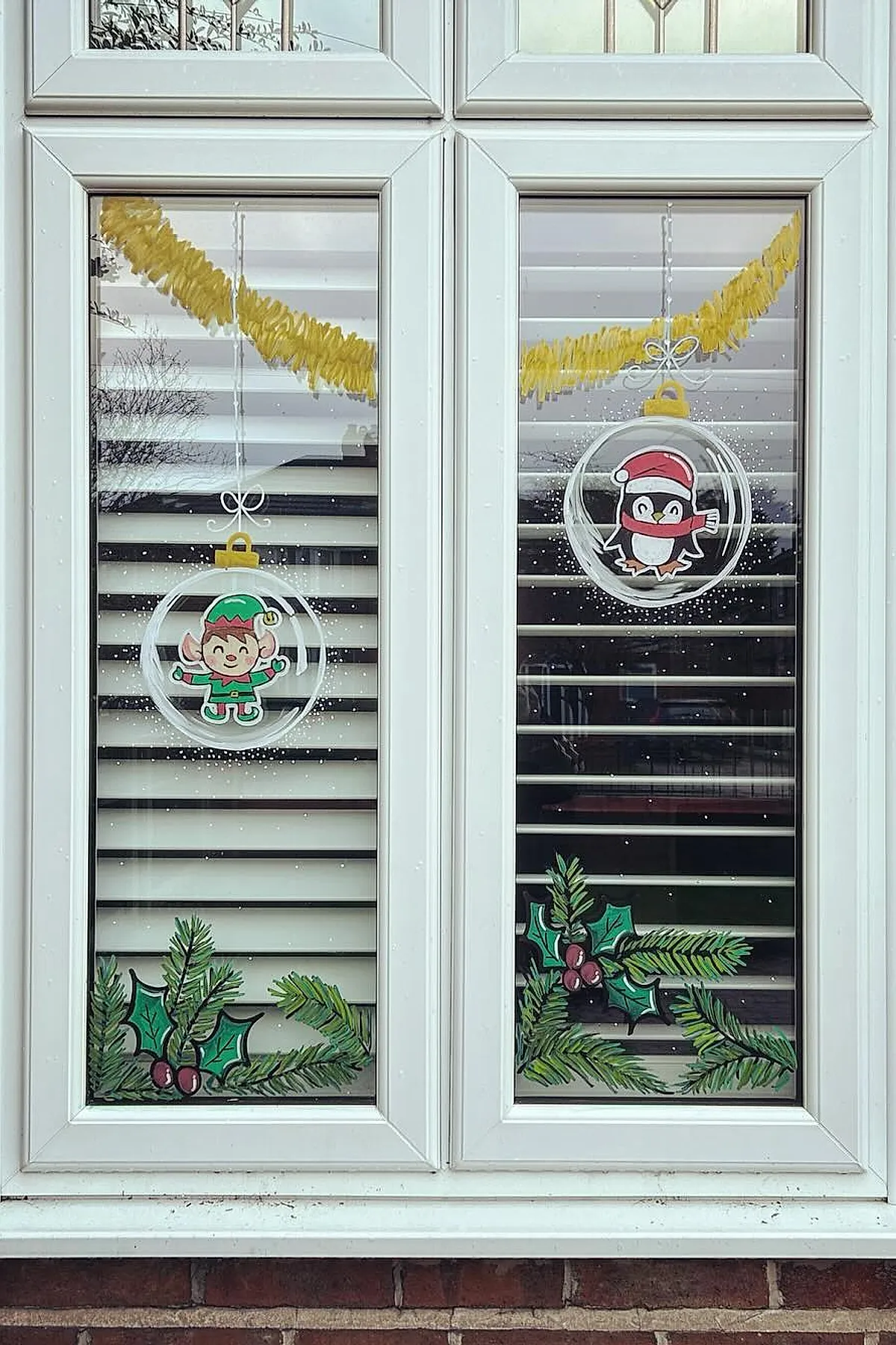 Christmas window painting showing two clear glass ornaments hanging from a yellow garland, one with a smiling elf and the other with a penguin in a red Santa hat, surrounded by painted holly leaves and berries along the bottom of the window.