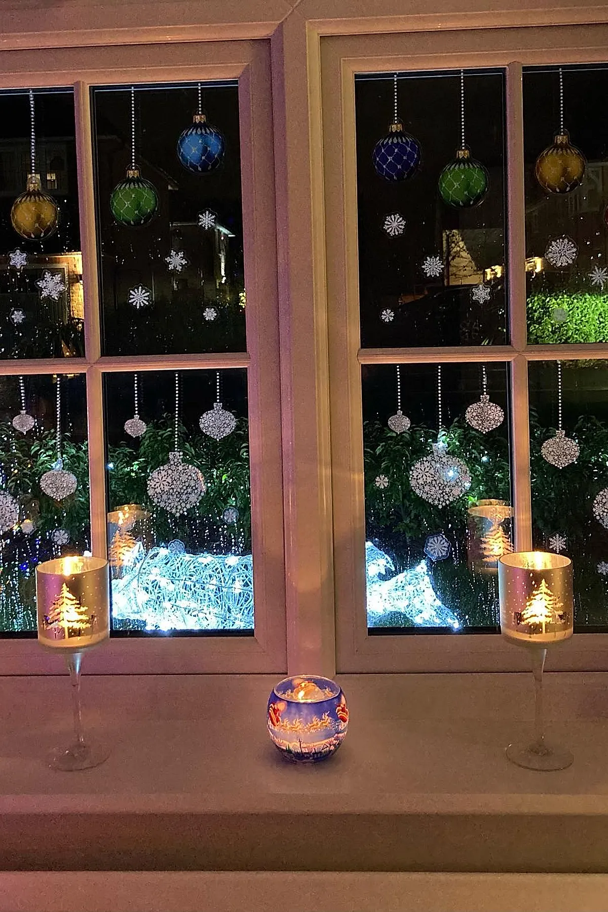 Holiday window art with colorful ornament and white snowflake stickers hanging in rows, paired with candle holders featuring tree cutouts and glowing candles on the sill for a cozy winter window display.