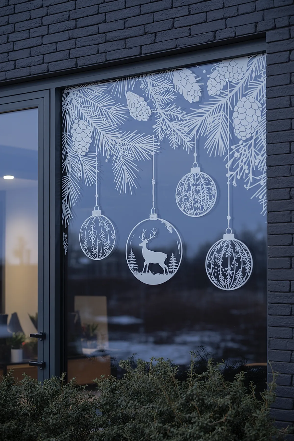 Christmas window art on a large glass pane featuring hanging ornaments, pine branches, and pinecones painted in white, creating an elegant and detailed winter scene on a dark frame.