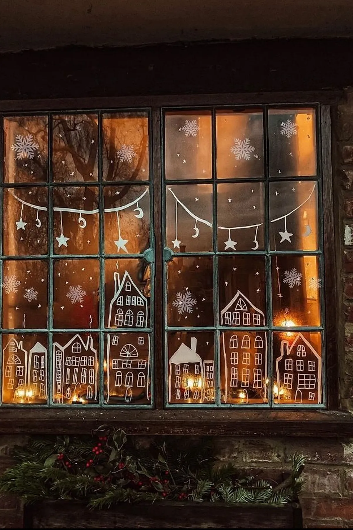 Christmas window painting depicting white outlines of houses along the bottom panes with hanging stars and crescent moons above, surrounded by snowflakes and accented with greenery and red berries on the windowsill.