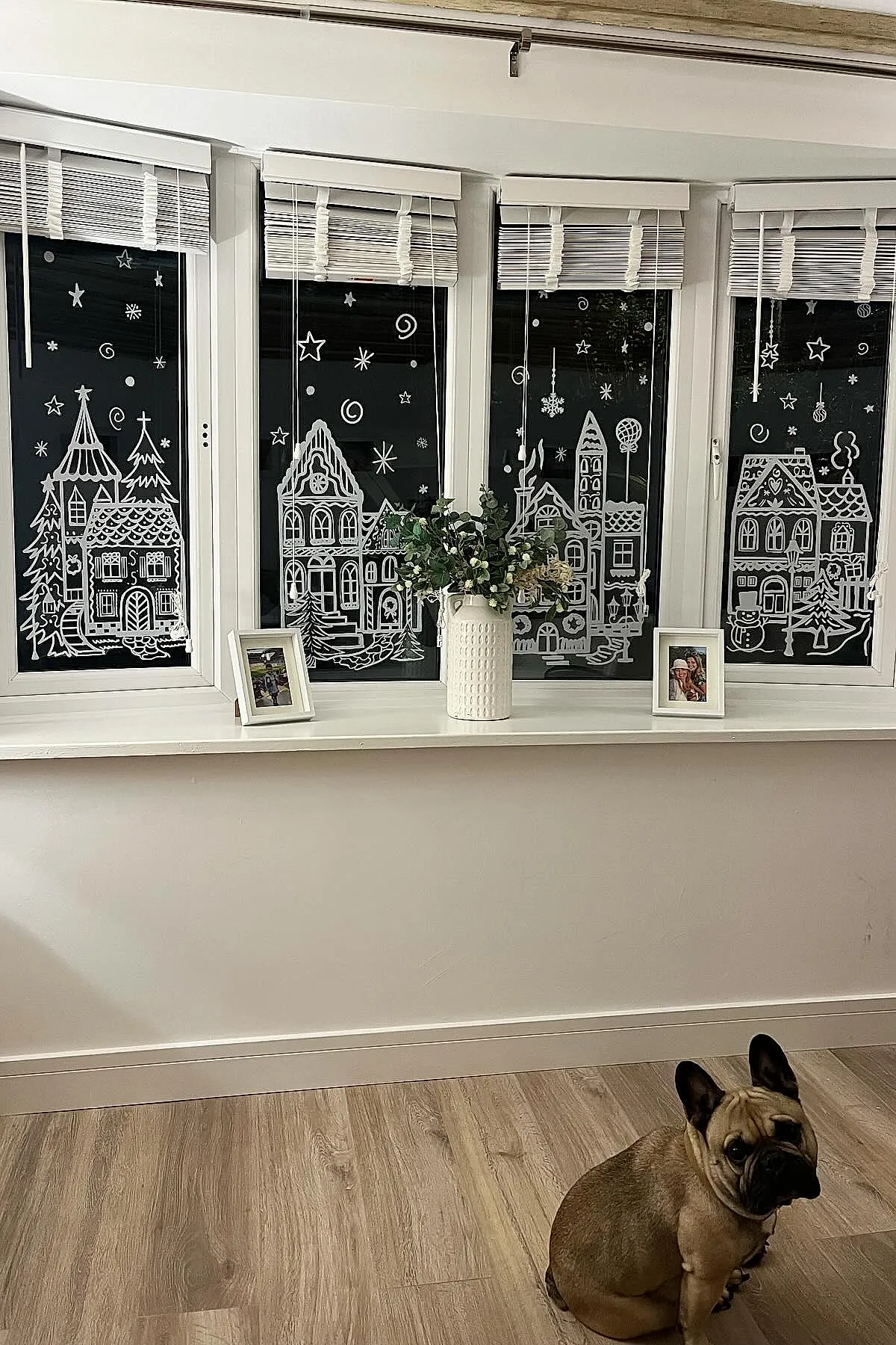 Christmas window art featuring a white-painted winter village scene with houses, trees, stars, and swirling snowflakes, accented with greenery in a vase on the sill for a storybook-style display.