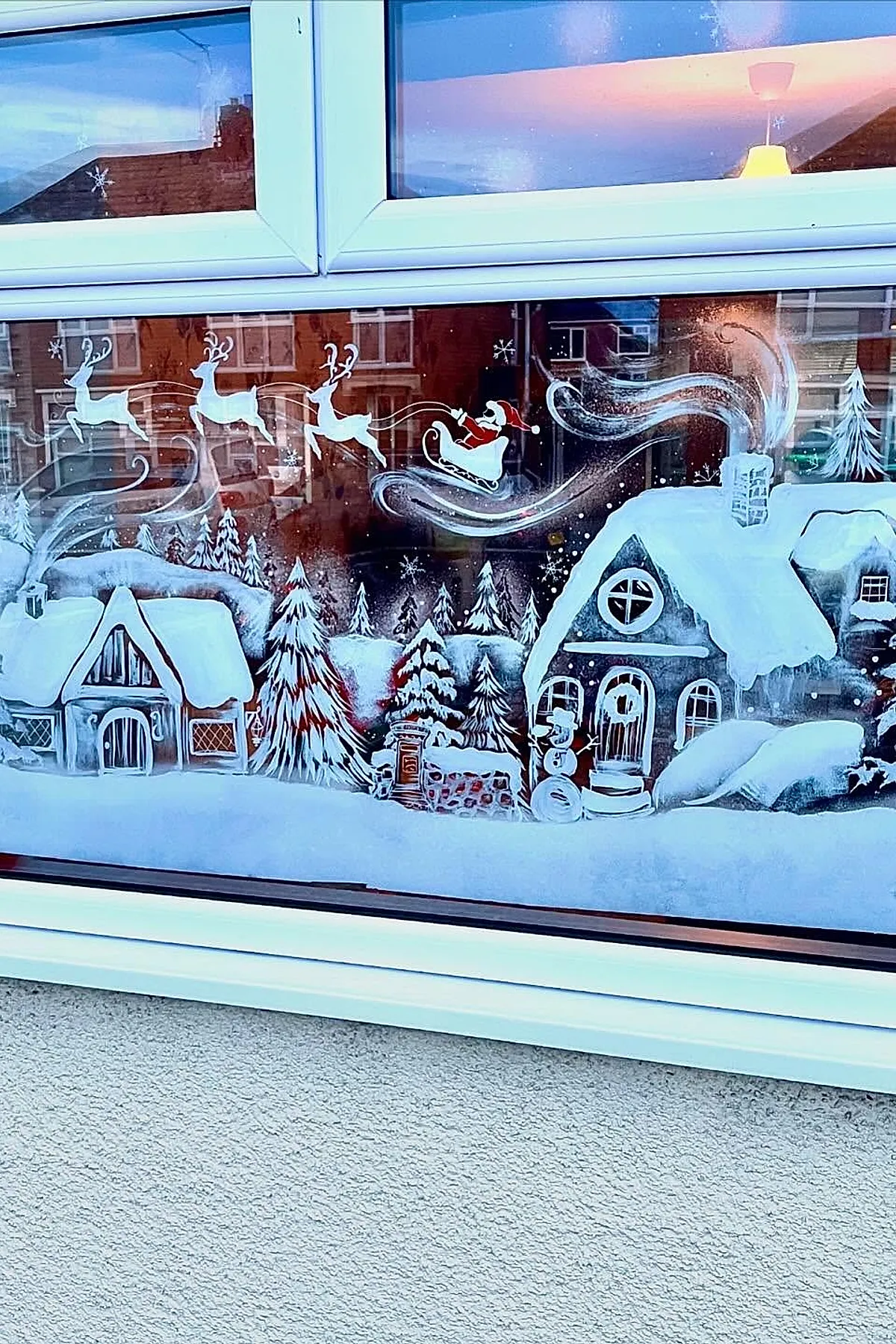 Christmas window painting showing Santa Claus in a red suit riding a white sleigh pulled by reindeer flying over a snowy village with rooftops, trees, and swirling chimney smoke for a classic holiday window art design.