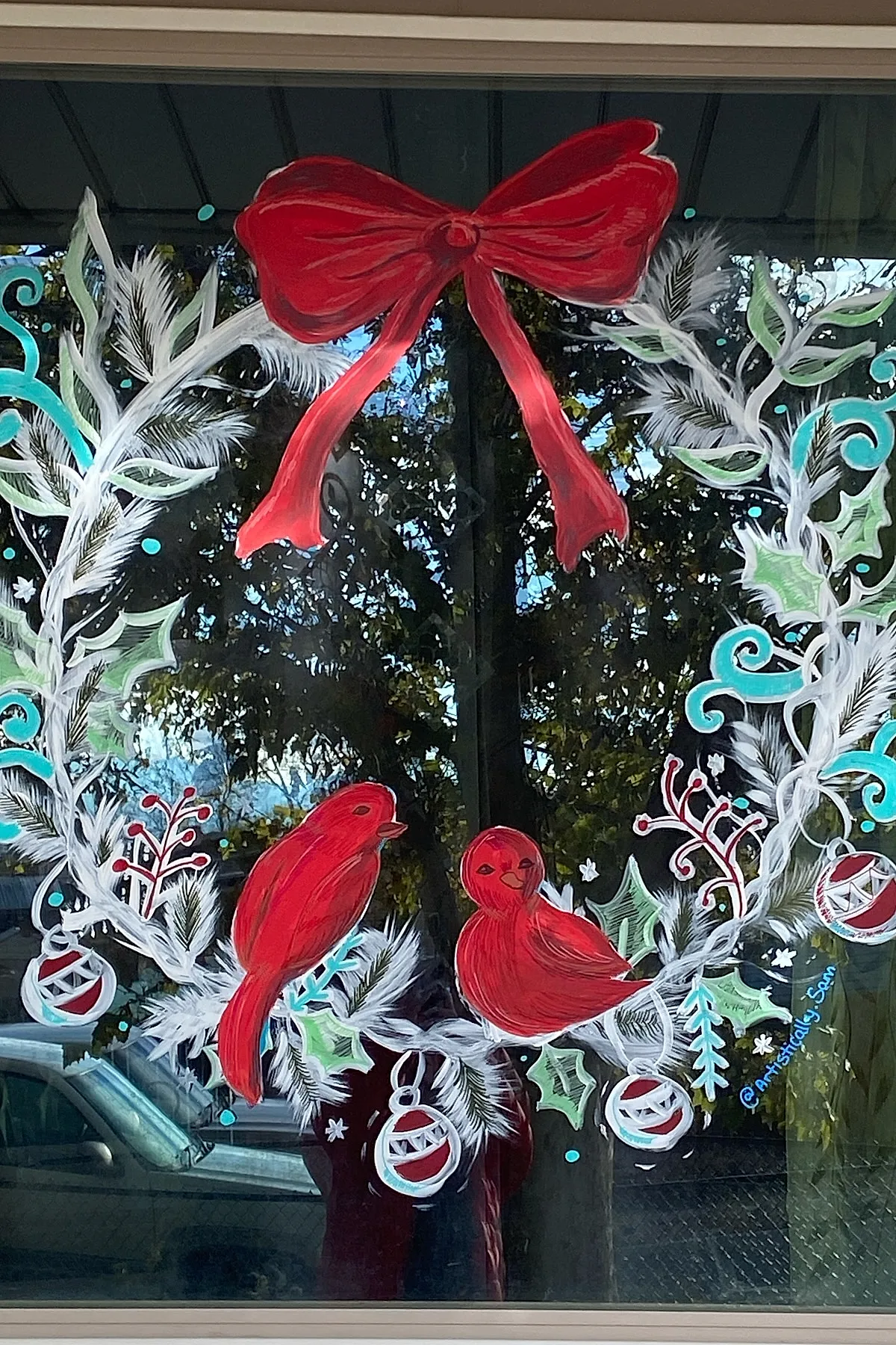 Christmas window art featuring a painted wreath with white feathery branches, green holly leaves, turquoise swirls, red berries, and two red birds perched inside for a festive holiday window idea.