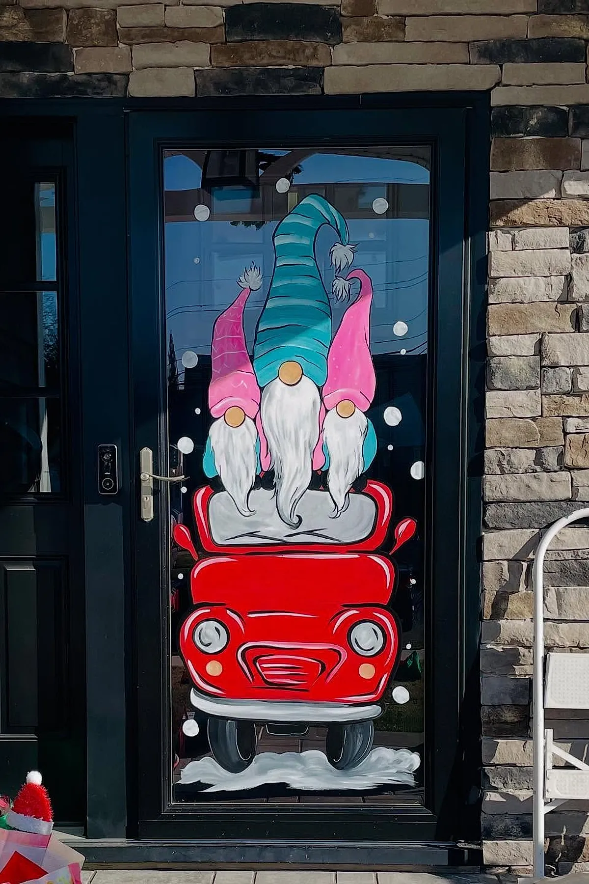 Christmas window painting featuring three gnomes with long white beards and striped blue and pink hats riding in a bright red car surrounded by white snowflakes, a cheerful holiday window art scene.