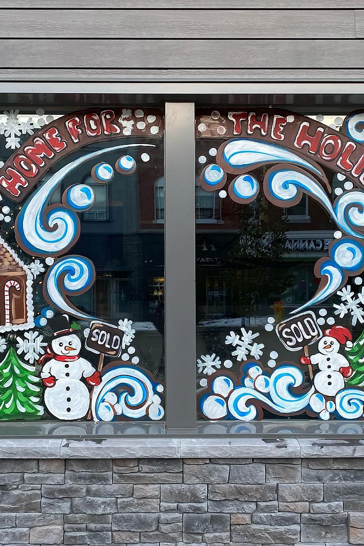 Christmas window painting showing two smiling snowmen holding 'sold' signs surrounded by swirling blue and white snow patterns, green trees, and a gingerbread house for a fun winter window display.