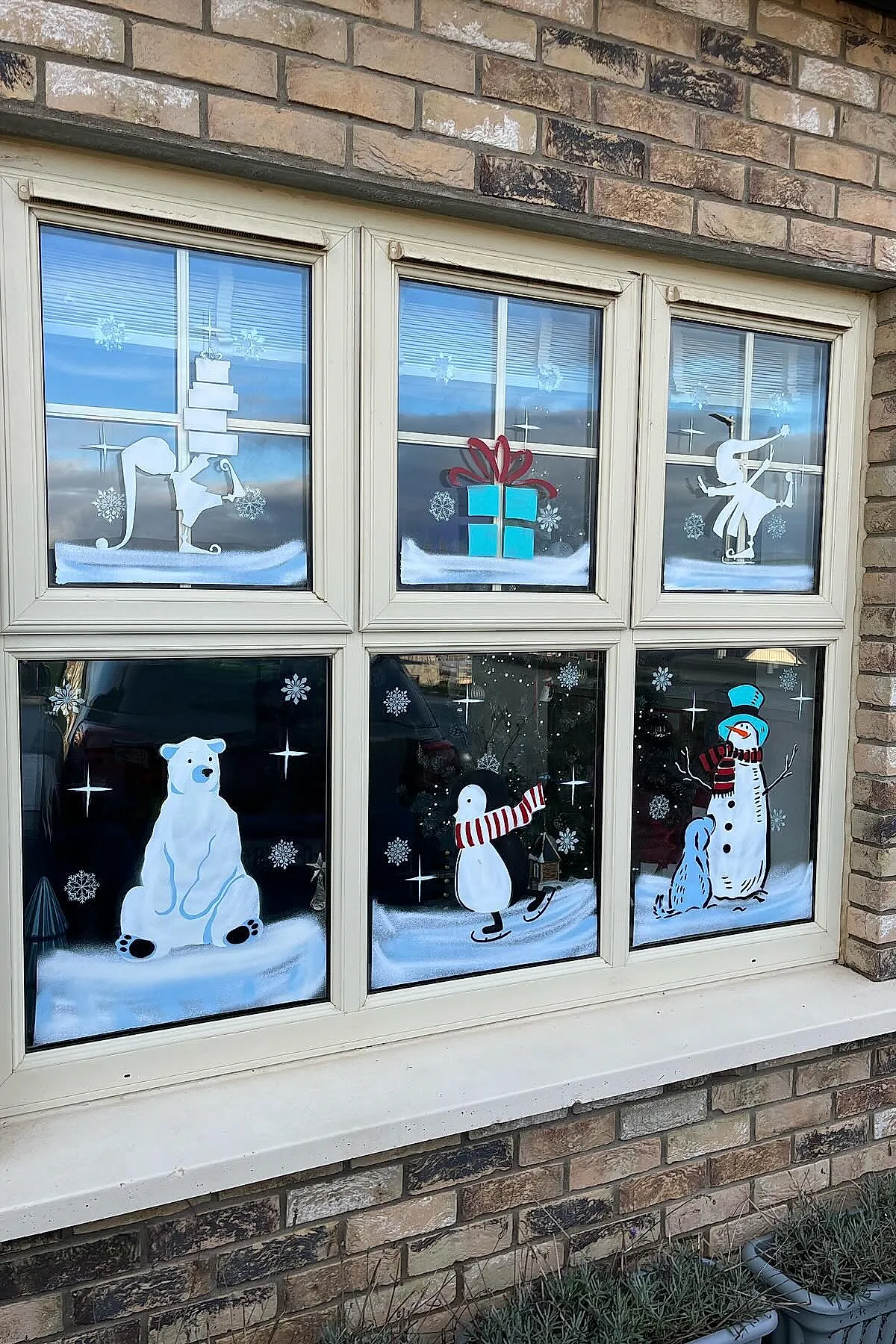 Christmas window painting featuring playful winter characters including elves with gifts, a polar bear, skating penguin, and snowman surrounded by white painted snowflakes and stars across multiple panes for a lively holiday window art design.