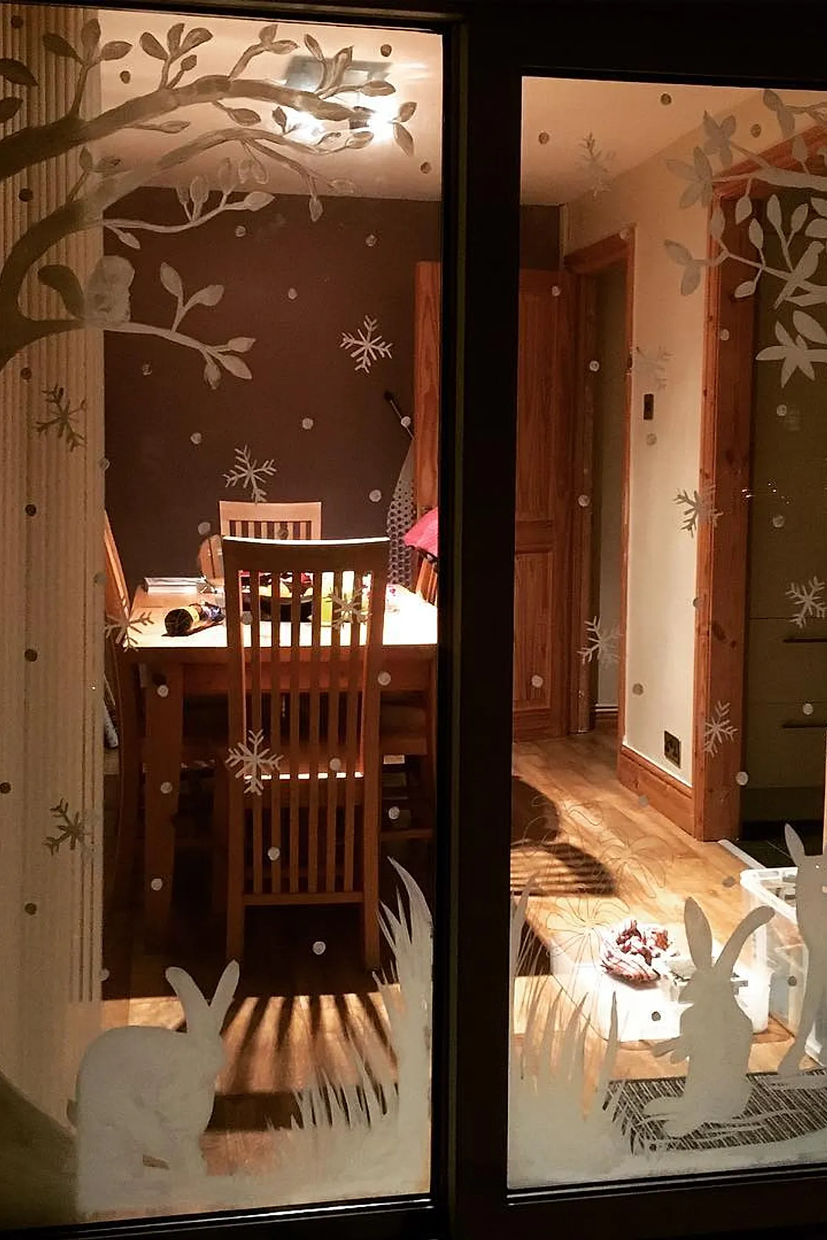 Christmas window art featuring white painted silhouettes of rabbits sitting in grass beneath large snowy tree branches with scattered snowflakes and dots, creating a serene winter window display.