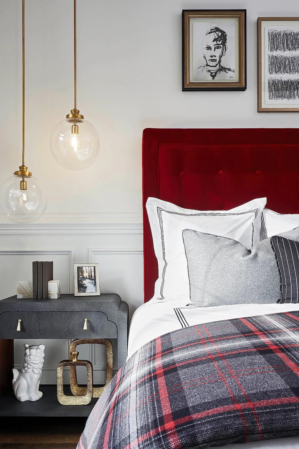 bedroom with red velvet tufted headboard, white bedding with black piping, gray and black pillows, red and gray plaid blanket, dark gray bedside table with decorative objects, two framed abstract artworks on white wall, hanging glass pendant lights