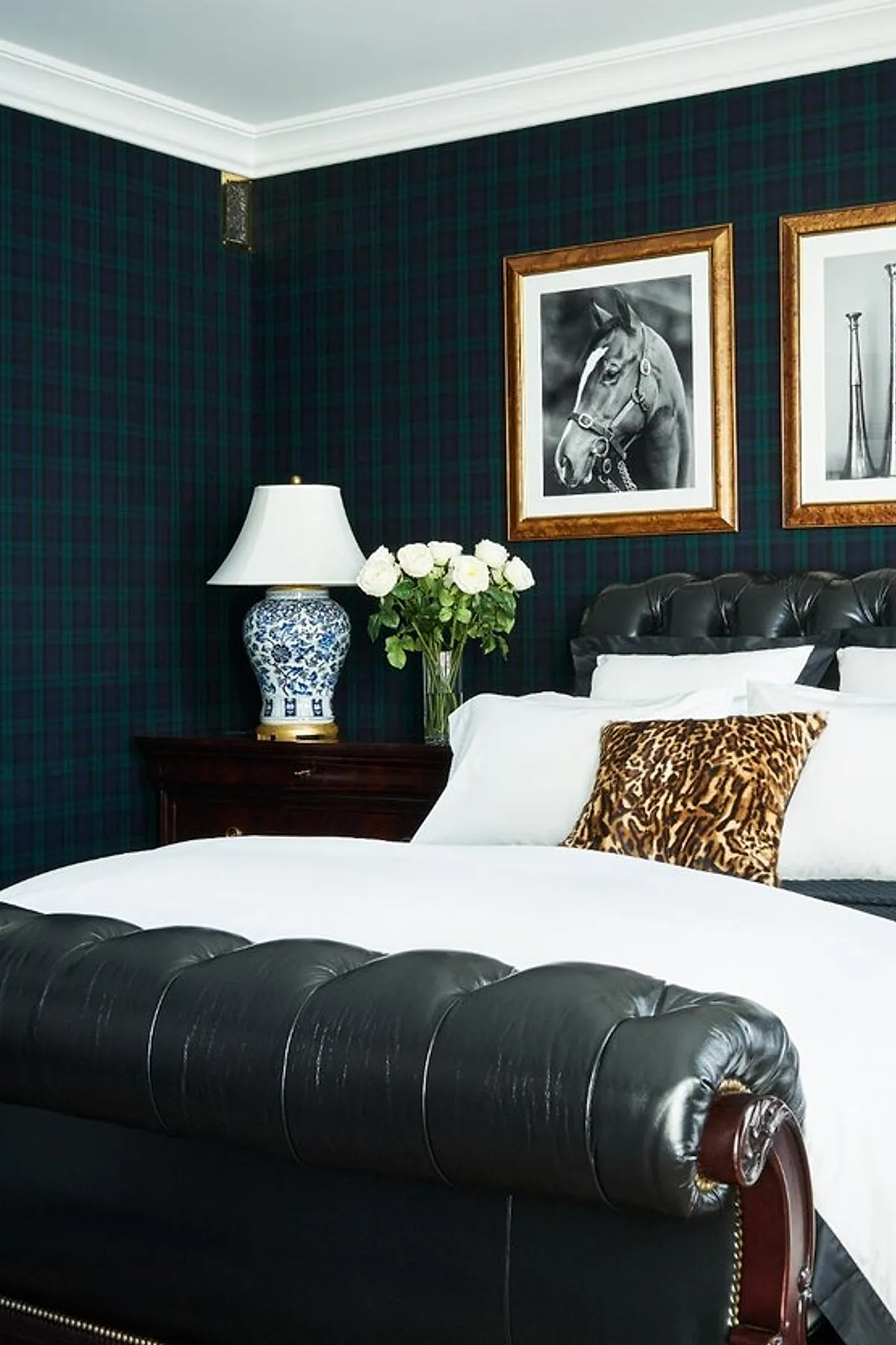 bedroom with dark green and navy plaid wallpaper, black leather tufted bed, white bedding, leopard print pillow, blue and white ceramic lamp, framed horse photograph on wall