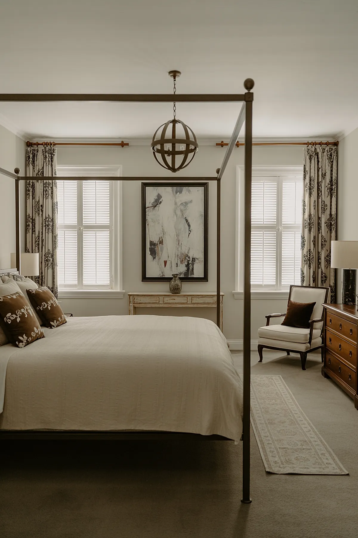 bedroom with neutral beige walls, metal four-poster bed with cream bedding, brown decorative pillows, patterned beige and black curtains on either side of two windows with white shutters, abstract painting above distressed wooden console table, upholstered armchair with dark wood arms and brown cushion