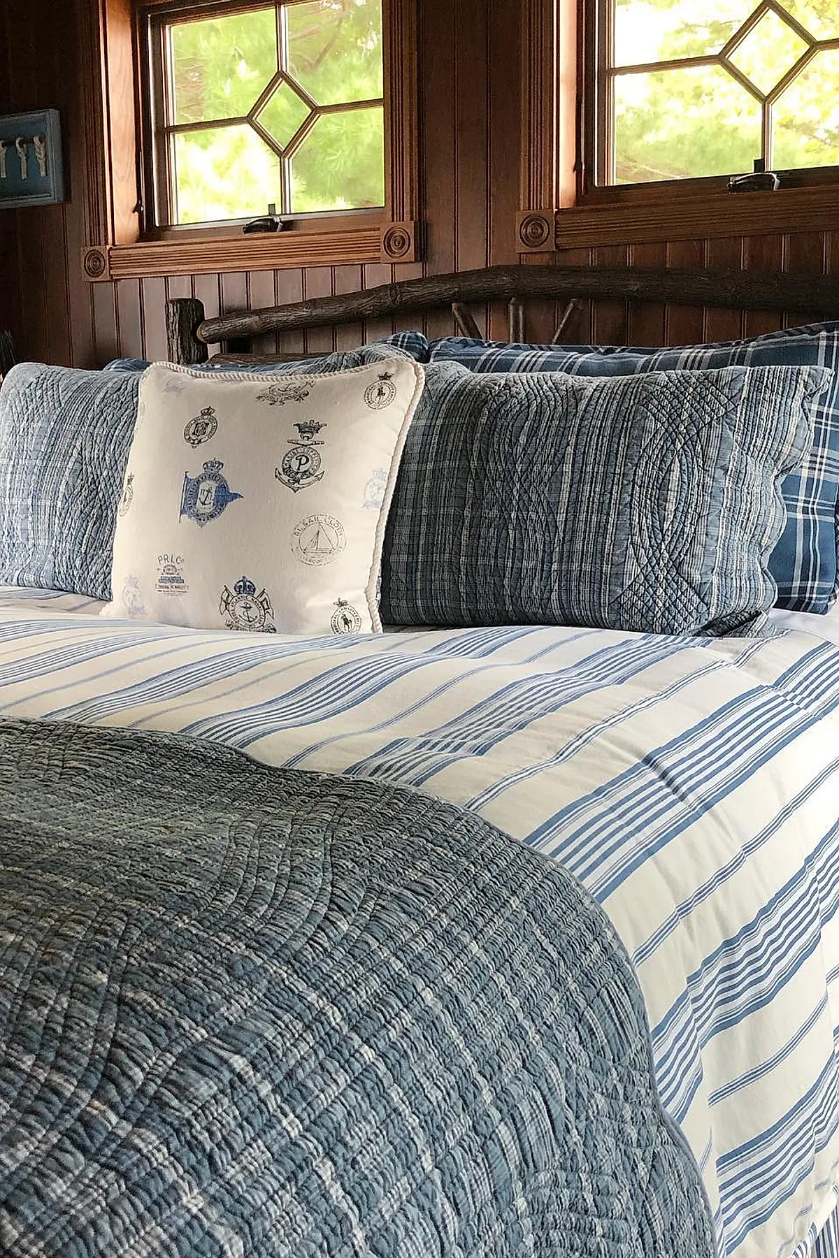 bed with blue and white striped bedding, quilted blue pillows, cream pillow embroidered with nautical crests, rustic dark wooden headboard against wood-paneled wall
