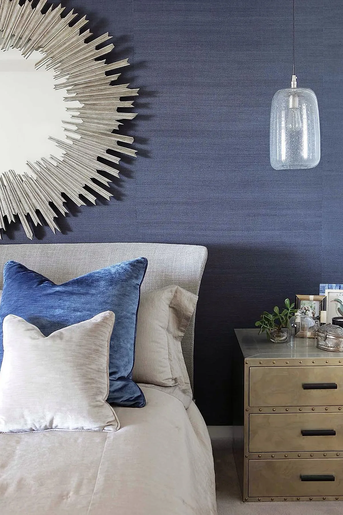 bedroom featuring textured navy blue grasscloth wall, silver sunburst mirror, light beige upholstered bed with blue velvet and cream pillows, metallic nightstand with black handles holding small plants and decorative items, clear glass pendant light