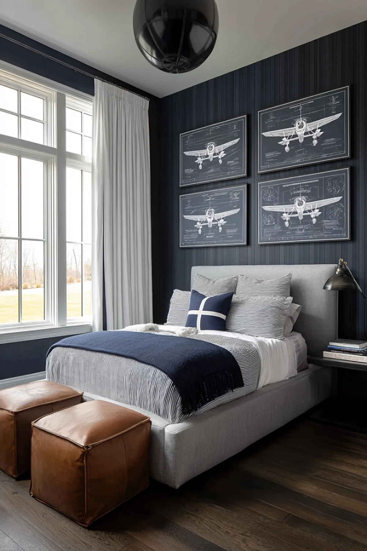 bedroom with dark navy striped walls featuring four framed airplane blueprint prints, gray upholstered bed with striped bedding and navy throw, two brown leather poufs at foot of bed, large white curtains over tall windows