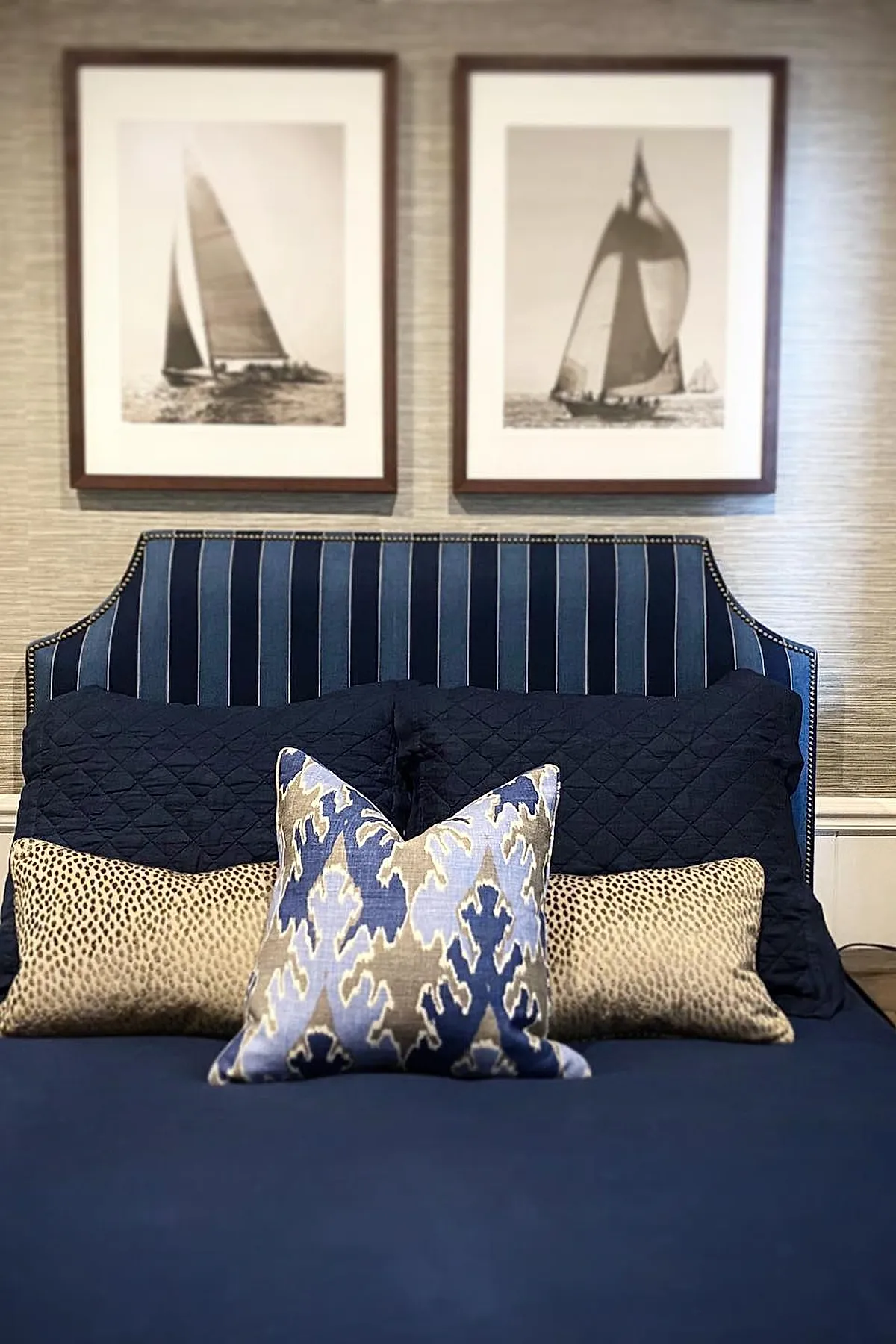 bed with navy and white striped upholstered headboard, navy quilted bedding, patterned blue and beige decorative pillows, two framed black and white sailboat photos above bed
