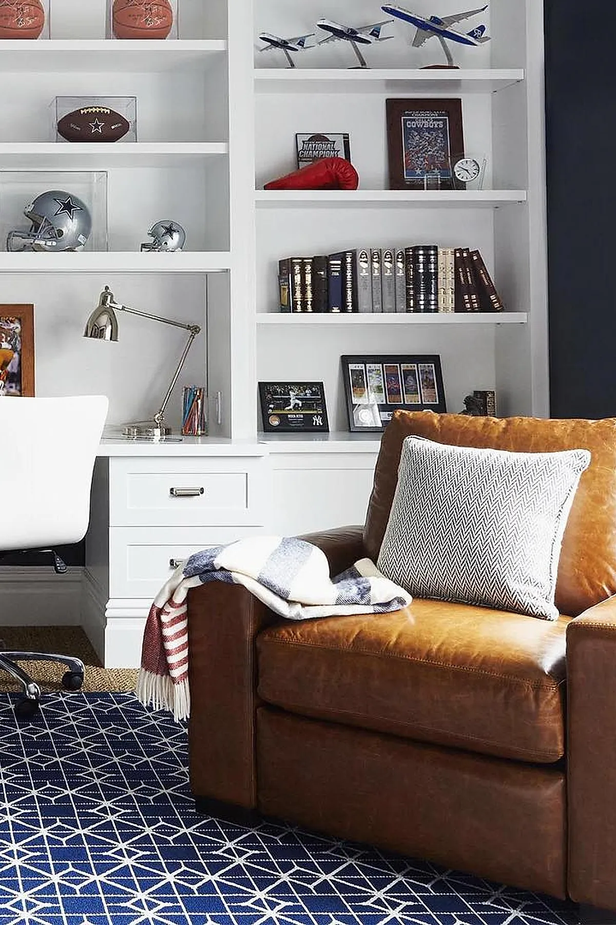 brown leather armchair with white patterned pillow and red white blue striped throw on navy blue geometric patterned rug in front of white built-in shelves filled with books model airplanes and sports memorabilia
