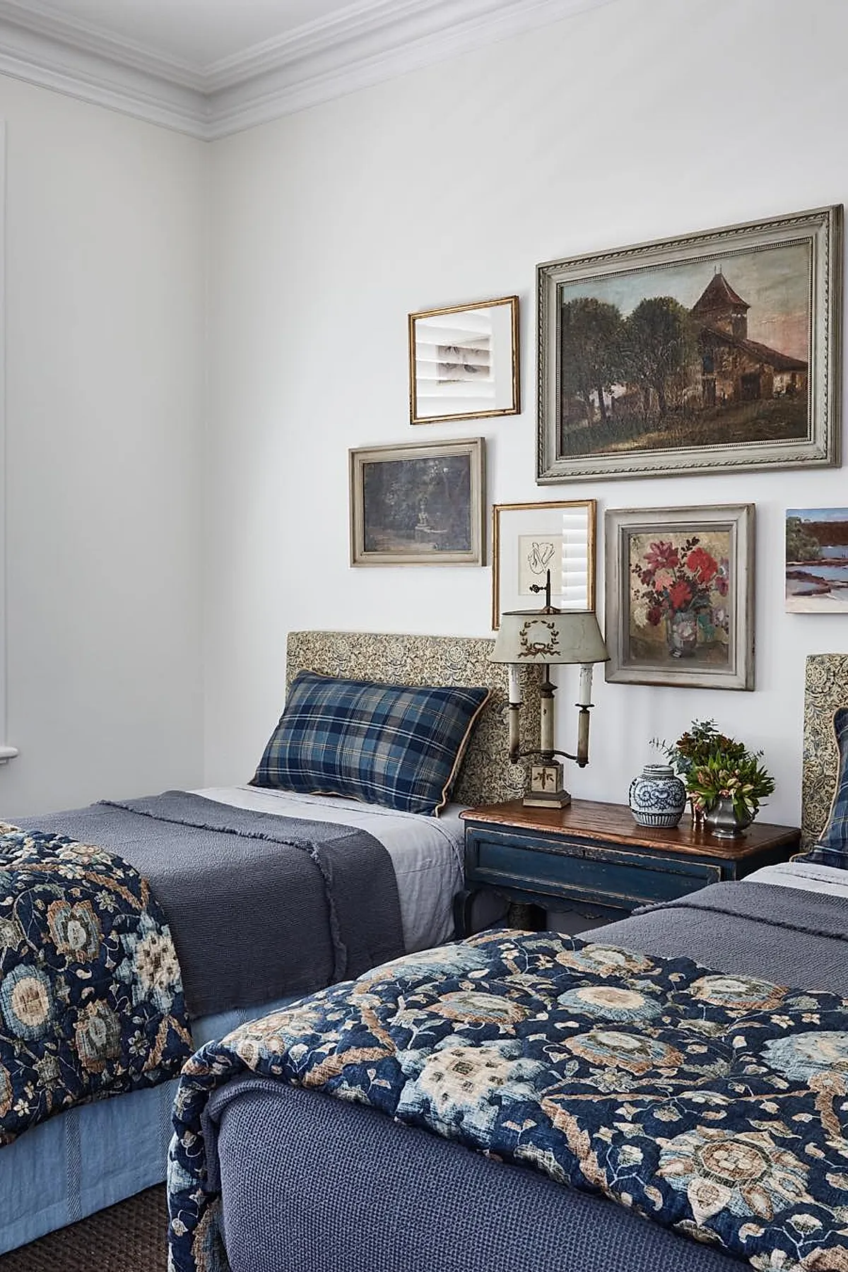 two twin beds with floral patterned headboards, blue plaid pillows, layered blue quilts and floral throws, vintage framed paintings and mirror arranged on white wall above wooden nightstand holding antique-style lamp and small plants